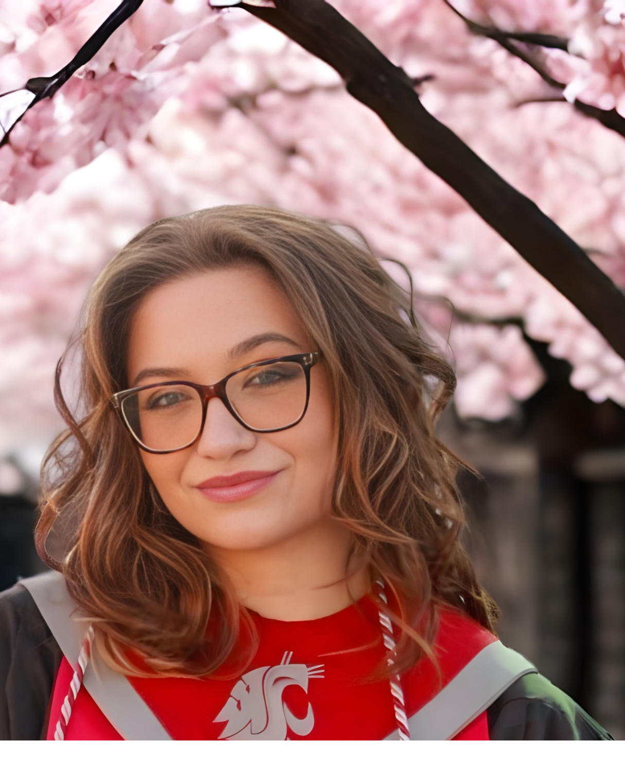 A young woman wearing glasses and a red graduation gown with a white logo, standing outdoors in front of pink cherry blossoms.