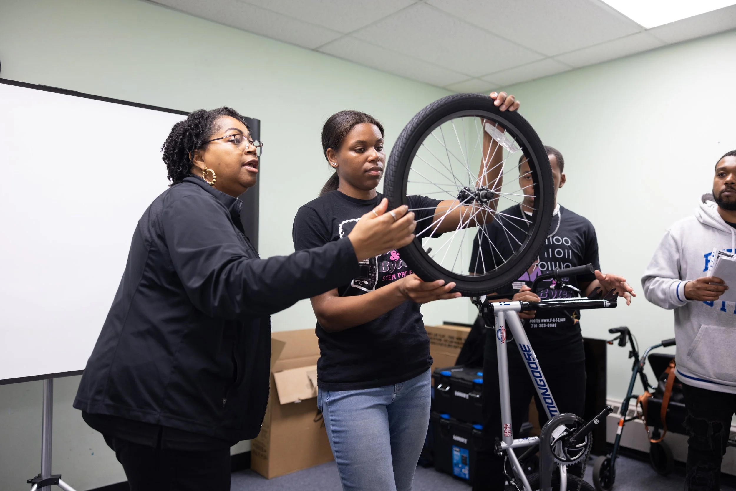 Build a Bike 5/22/25 — An image of a person teaching a teen on how to place a bicycle wheel onto a bike.