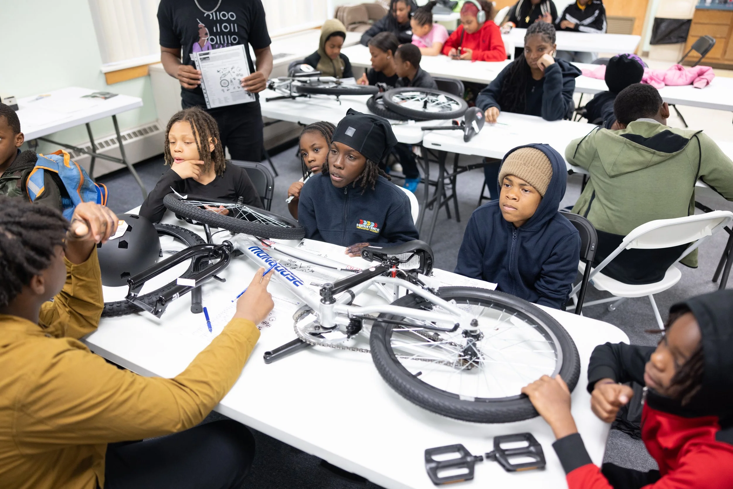 Build a Bike 5/22/25 — An image of teens sitting at a table learning about the different parts to a bike.