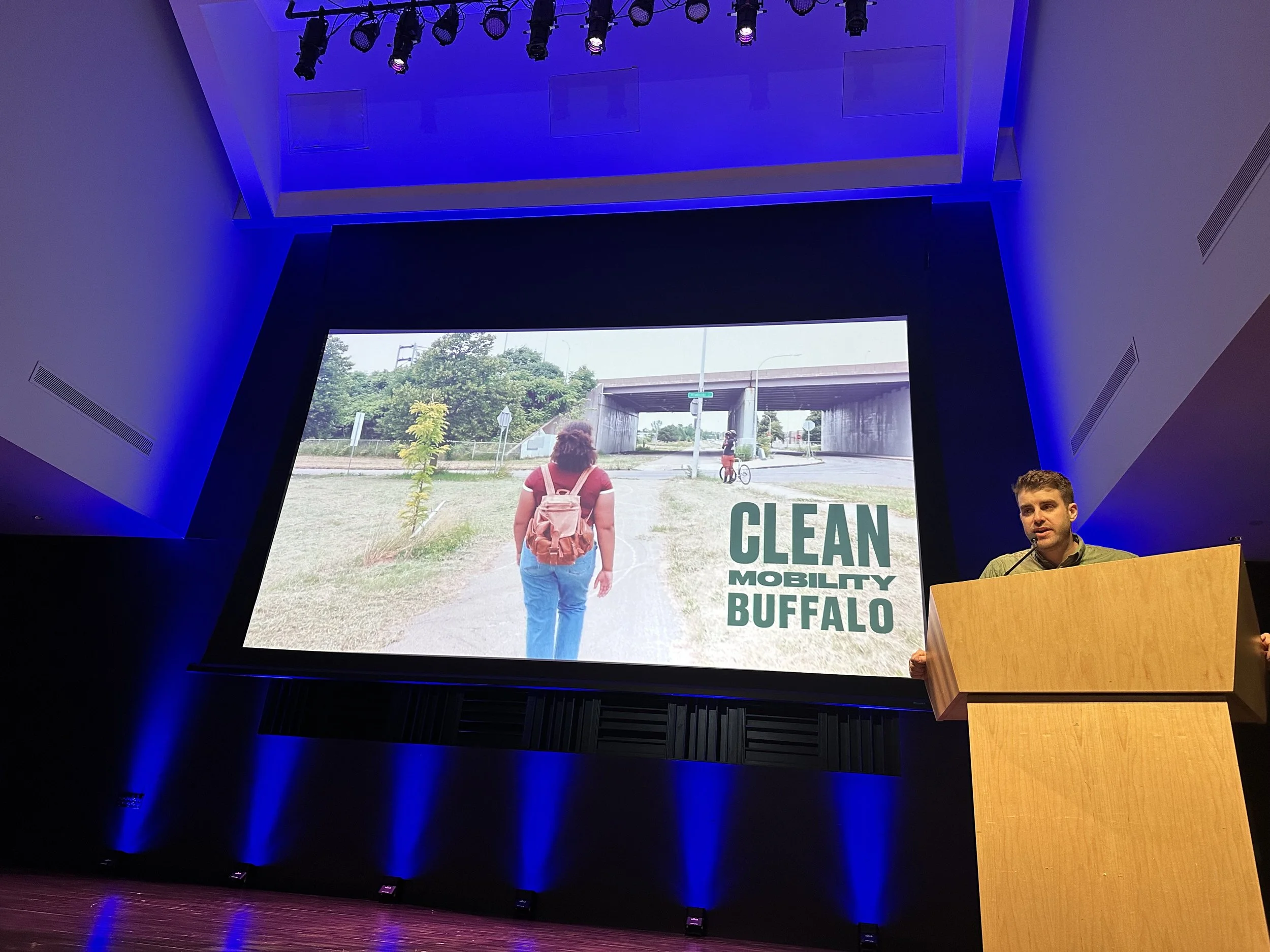Neighborhood Leadership Group at Ride Reels — An image someone on a stage behind a podium with a screen behind them showcasing a Clean Mobility Buffalo video.