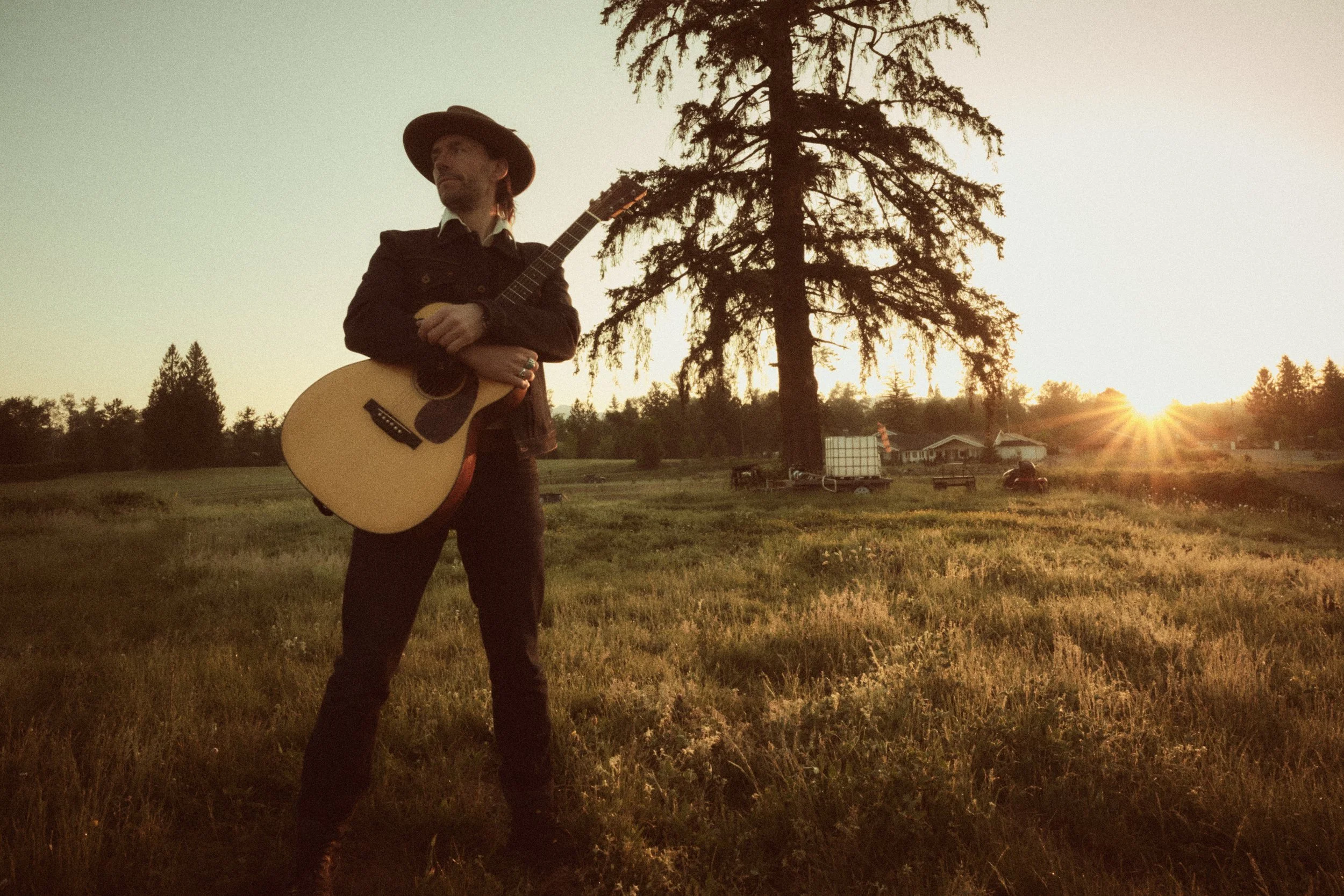 A man in a hat holding an acoustic guitar outdoors during sunset, with a large tree and houses in the background.