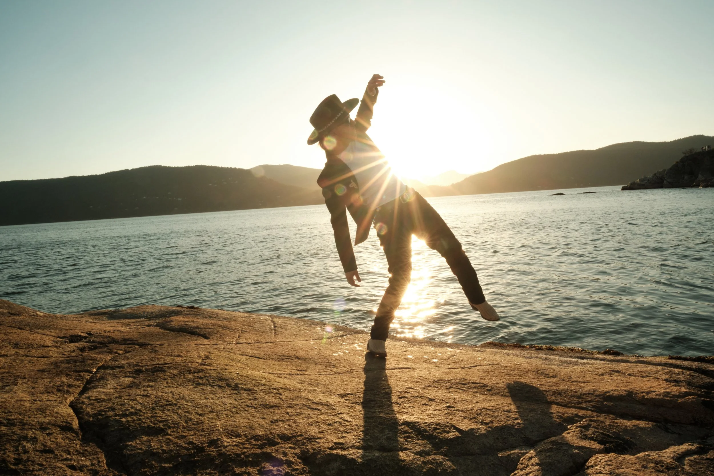 Person doing a cartwheel on rocks near a body of water during sunset, with hills in the background.