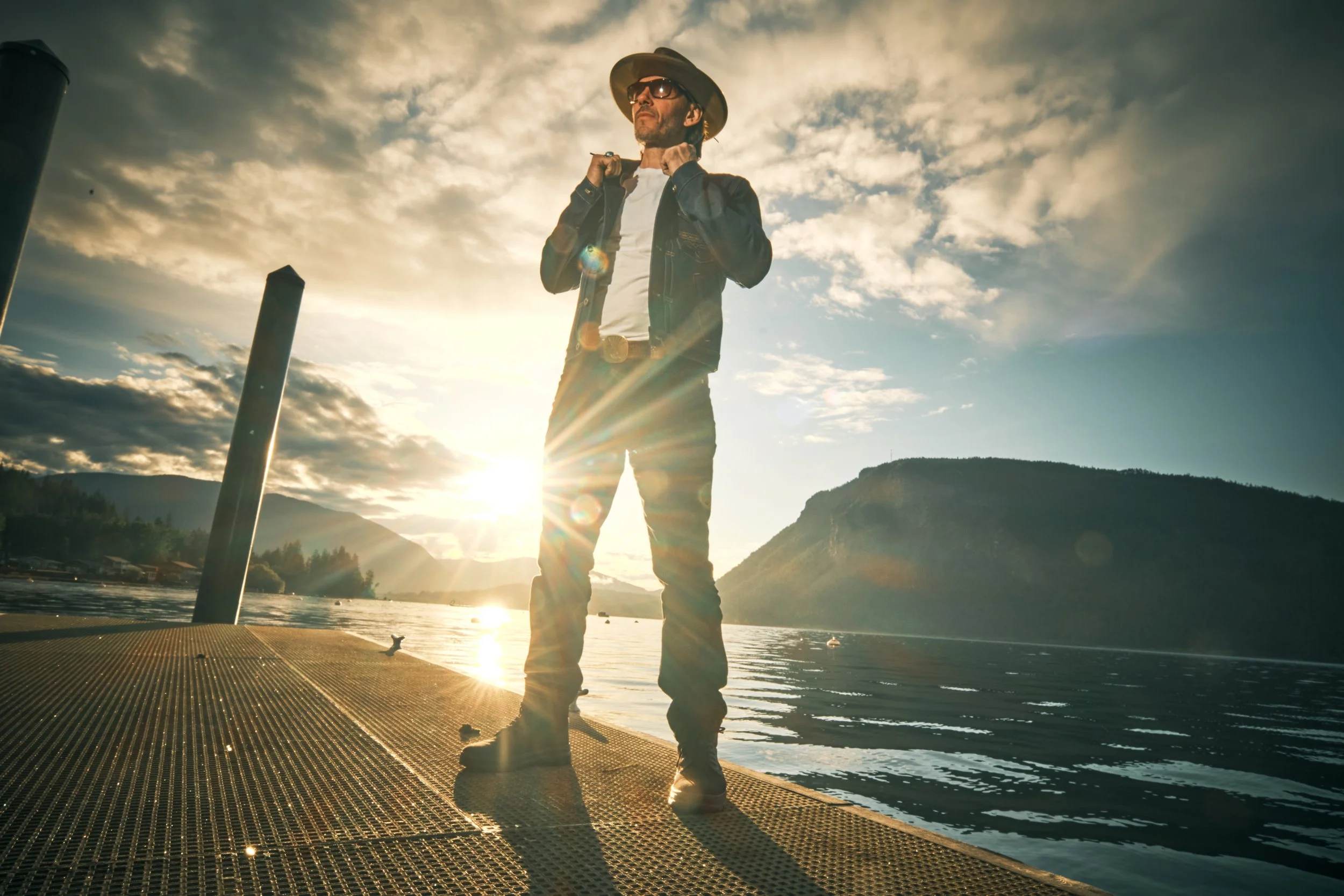A man standing on a dock near the water at sunset, wearing a hat, sunglasses, leather jacket, and jeans, with mountains in the background and the sun shining brightly behind him.