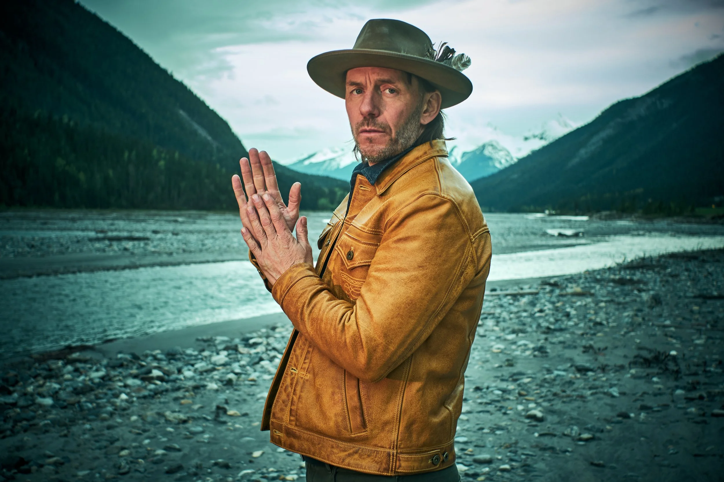 A man wearing a wide-brimmed hat and a tan jacket standing by a river with mountains in the background.