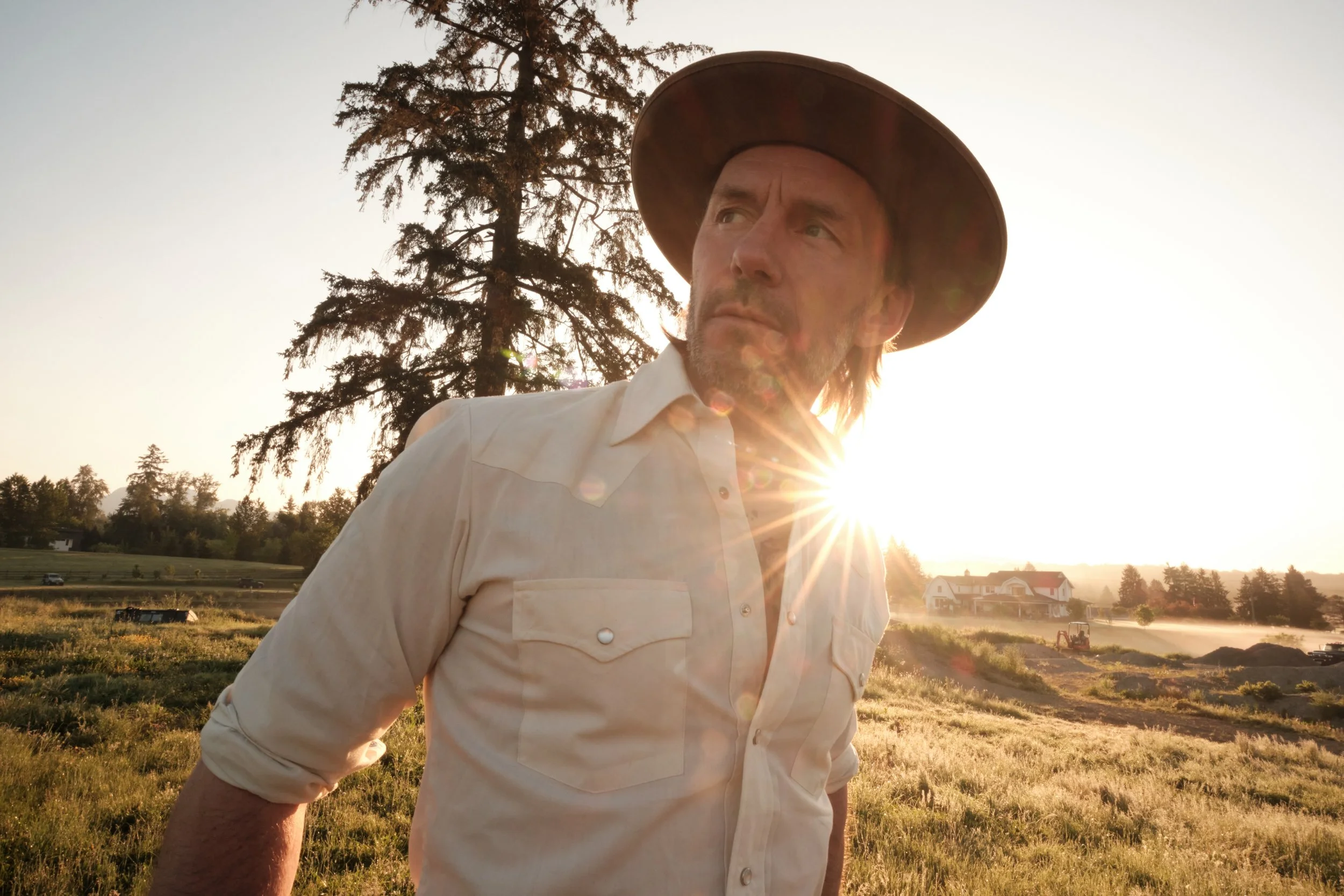 A man wearing a wide-brimmed hat and a light-colored shirt standing outdoors during sunset or sunrise, with a tree and houses in the background.