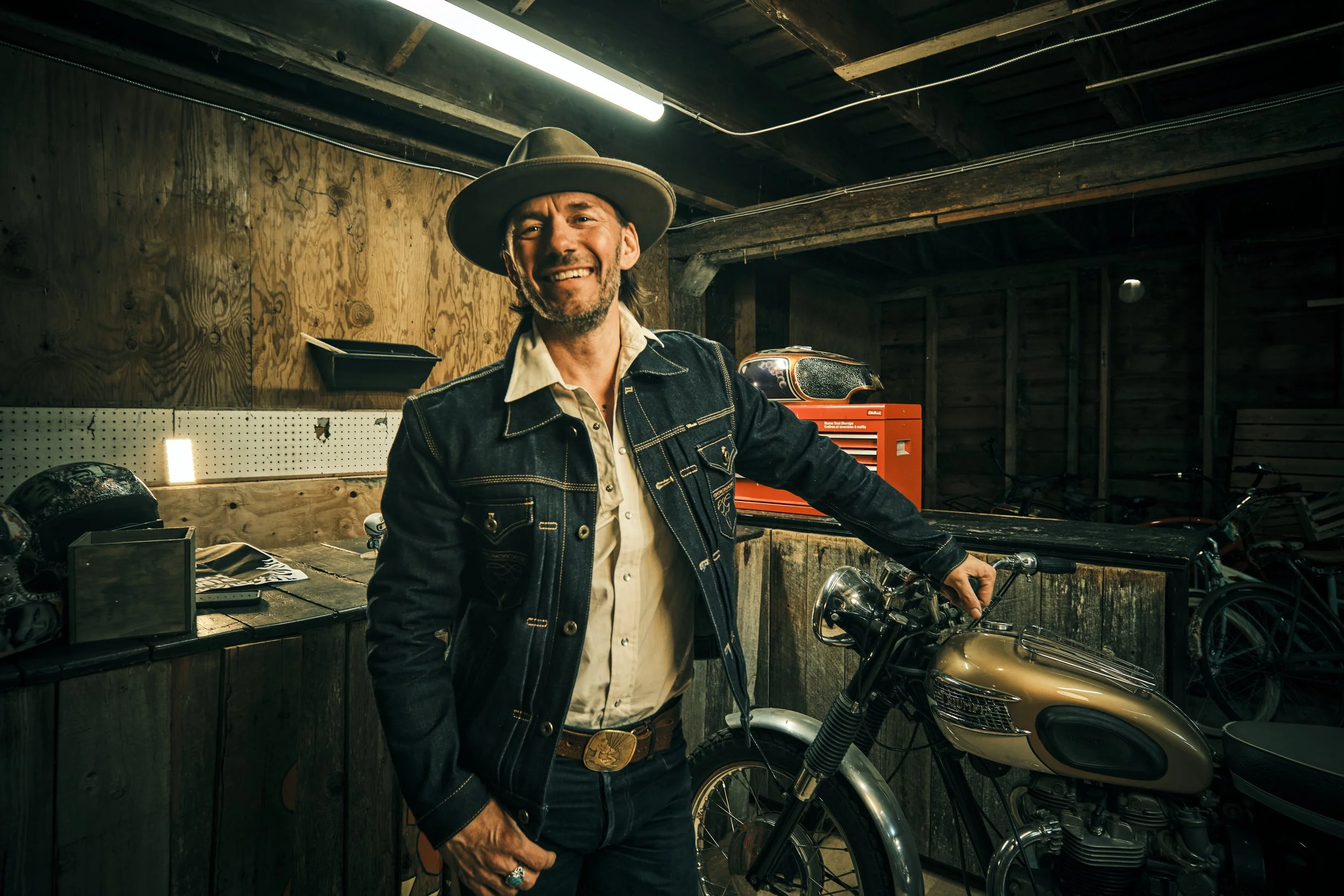 A man with long hair and a beard, wearing a wide-brimmed hat and denim jacket, standing in a garage with motorcycles and tools, smiling at the camera.