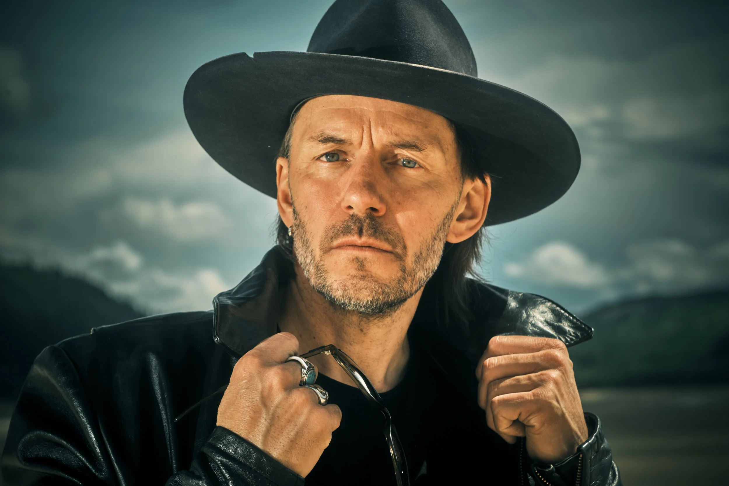 A man with gray facial hair wearing a wide-brimmed black hat and a black leather jacket standing outdoors against a cloudy sky.