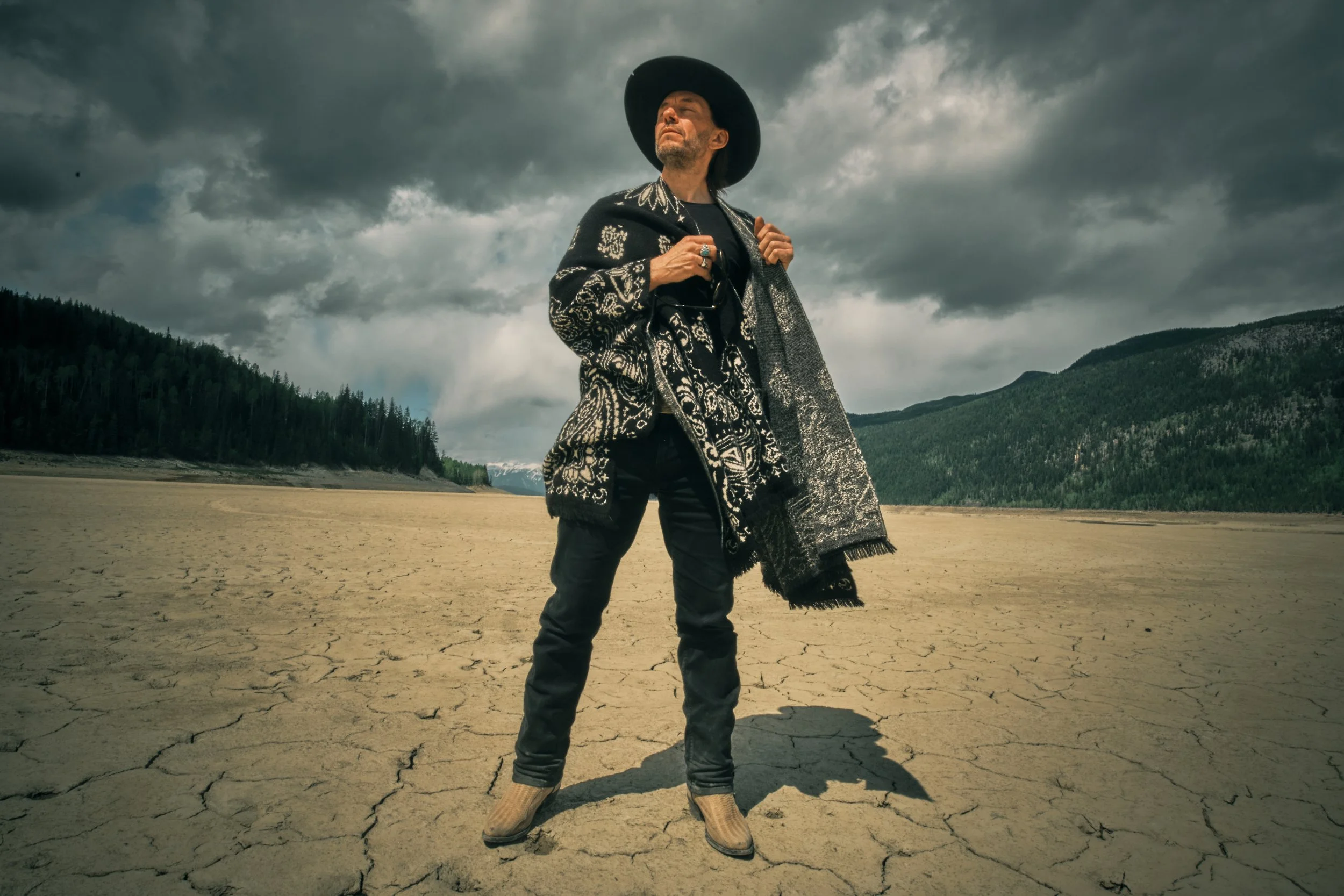 A person standing on a cracked dry lake bed under a dark, cloudy sky, wearing a large black hat, patterned jacket, black pants, and cowboy boots, holding glasses in one hand.