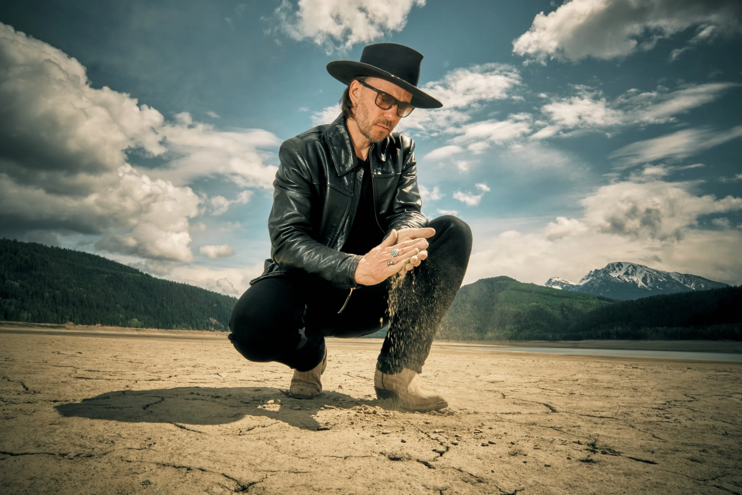 A man wearing a black leather jacket, sunglasses, rings, and a large black hat is kneeling on a cracked, sandy surface holding a handful of sand or dirt with a scenic mountain landscape and cloudy sky in the background.