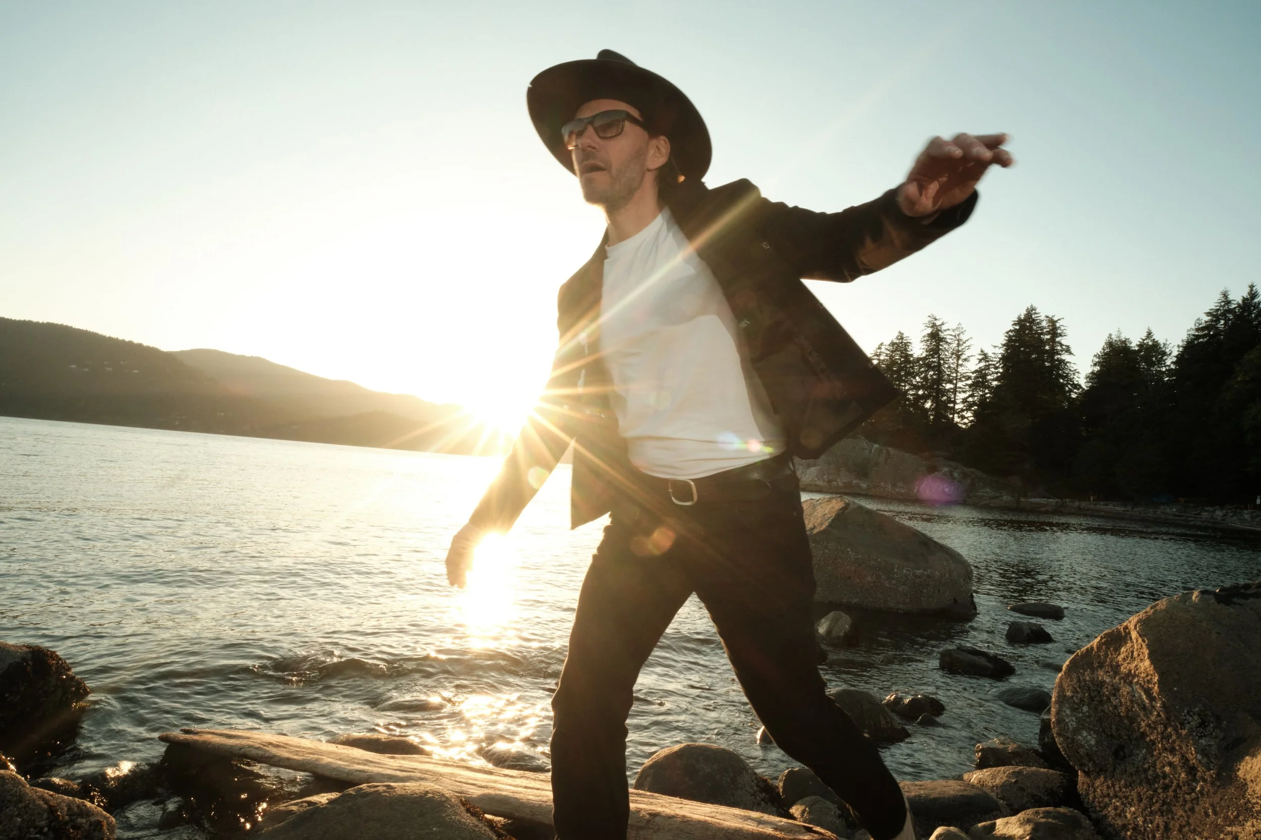 Man wearing hat and sunglasses standing on rocks at the edge of a body of water during sunset.