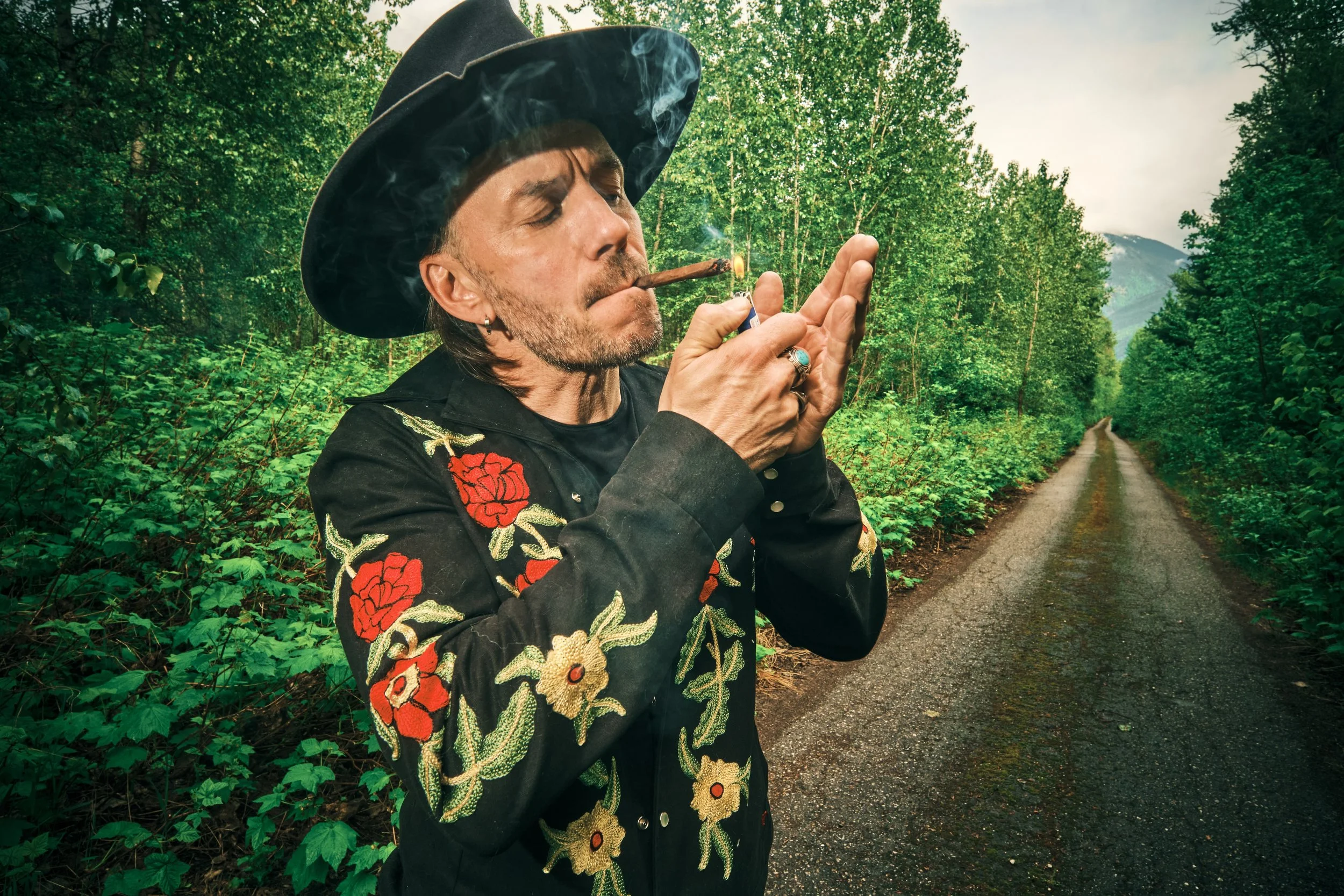 A man with long hair, wearing a black hat and black embroidered jacket, is lighting a cigar with a lighter on a dirt road surrounded by lush green trees in a forest.