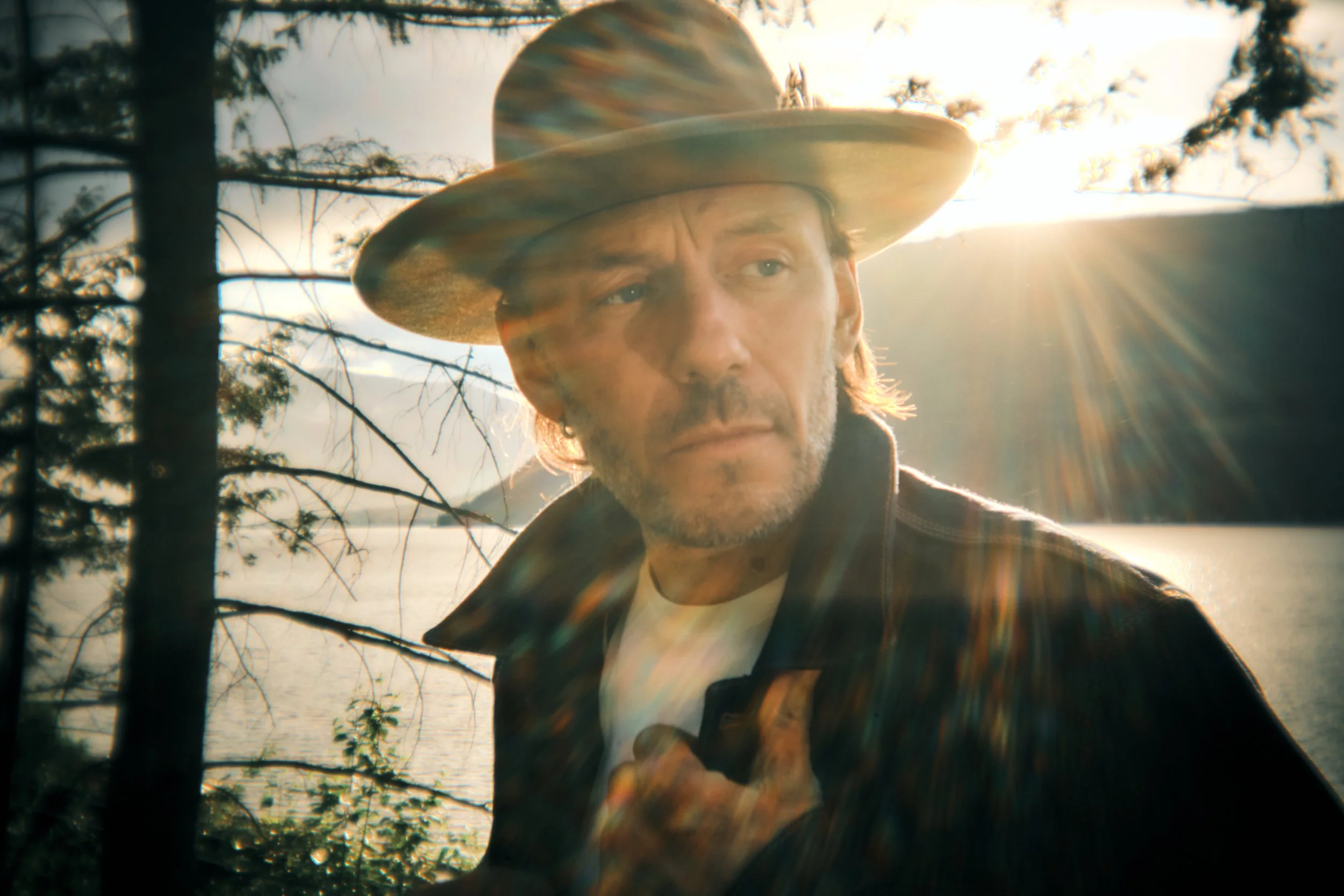 A man wearing a wide-brimmed hat standing outdoors near a body of water with trees, during sunset or sunrise, with sunlight creating lens flare effects.