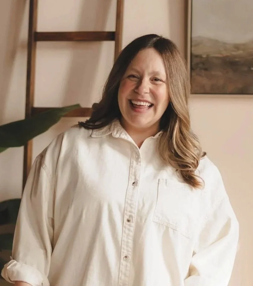 A portrait of Katie Wade showing a woman with wavy, shoulder-length brown hair,  with a big welcoming smile. She is wearing a casual but professional shirt.
