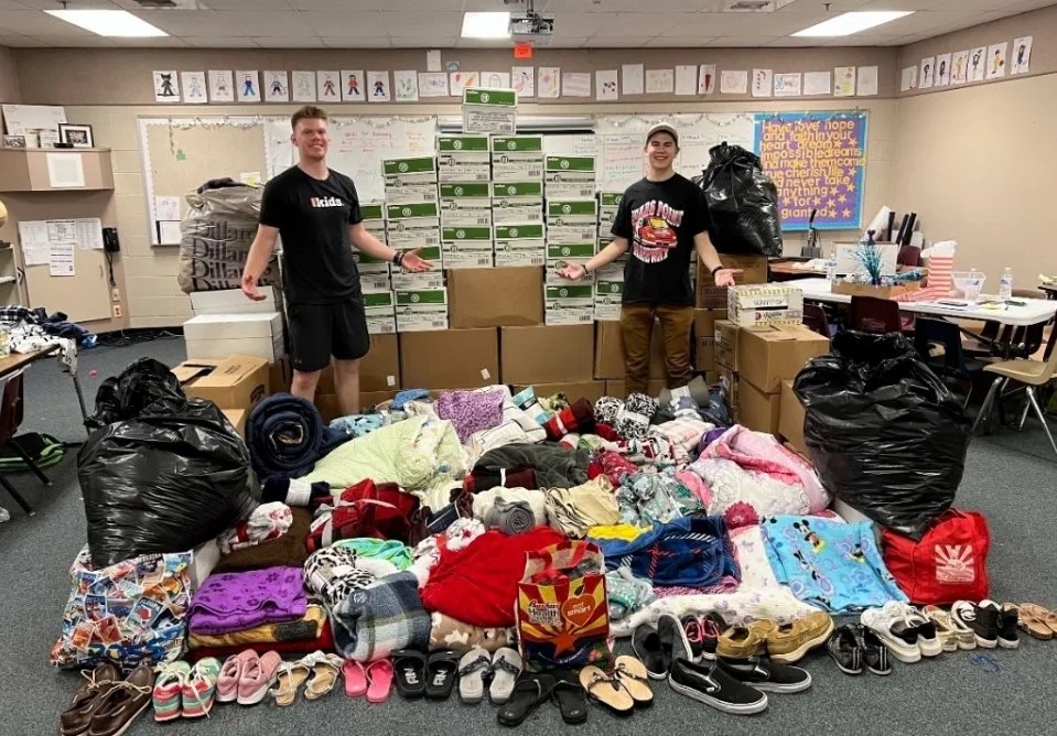 Two young men standing behind a large pile of donated clothing, shoes, and bags in a classroom with boxes and posters on the walls.