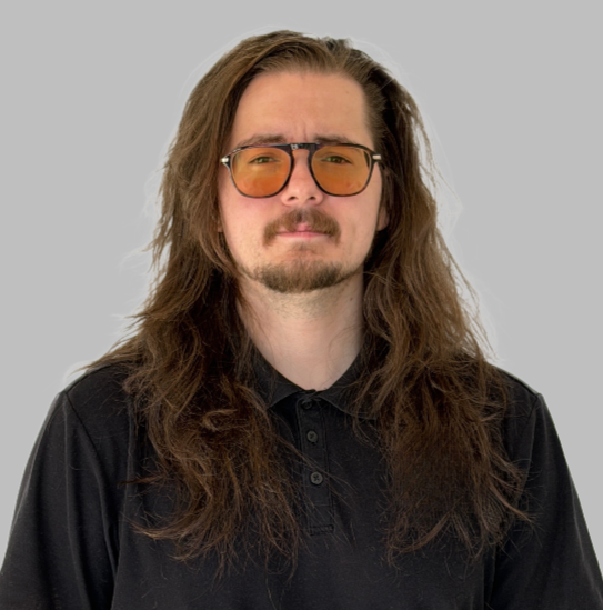 Man with long brown hair, wearing orange-tinted glasses and a black polo shirt, standing against a gray background.