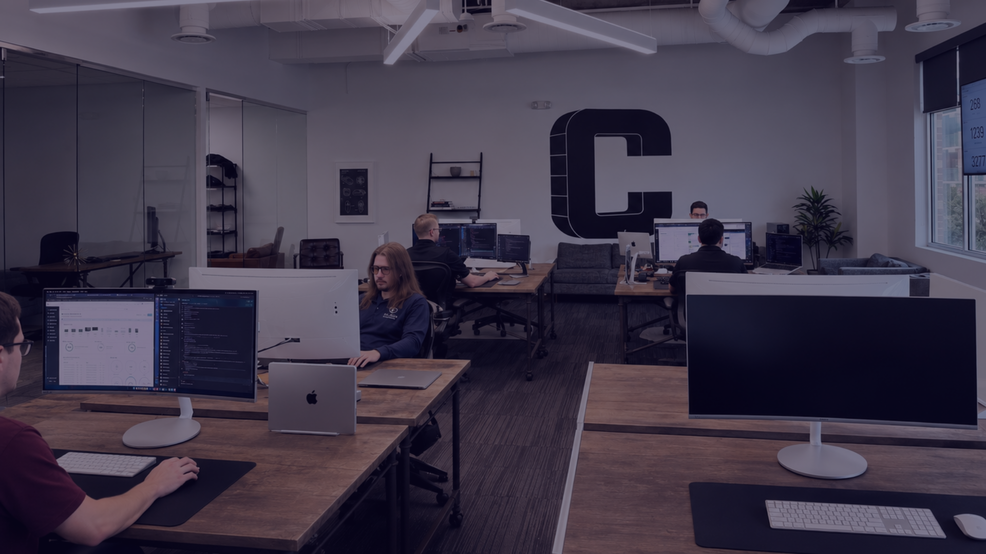 A modern office space with employees working at desks with multiple monitors, a large logo with the letter 'C' on the wall, and large windows letting in natural light.