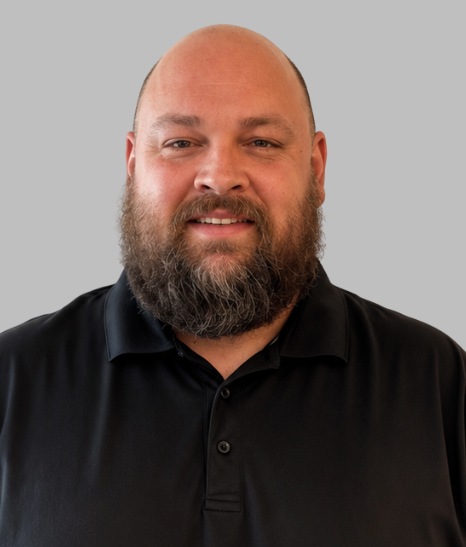 A bald man with a beard wearing a black collared shirt, looking at the camera, against a plain gray background.