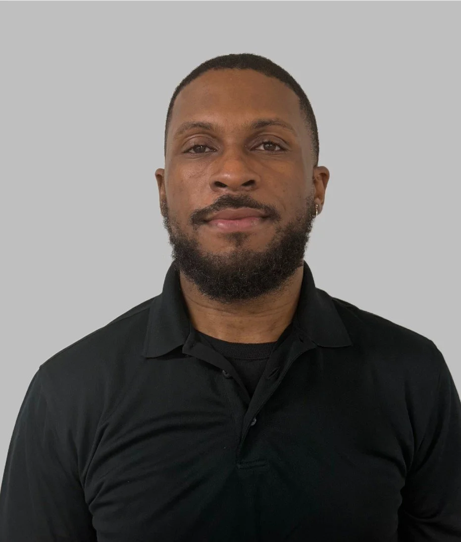 A man with short hair and a beard wearing a black collared shirt, facing the camera with a neutral expression, against a plain gray background.