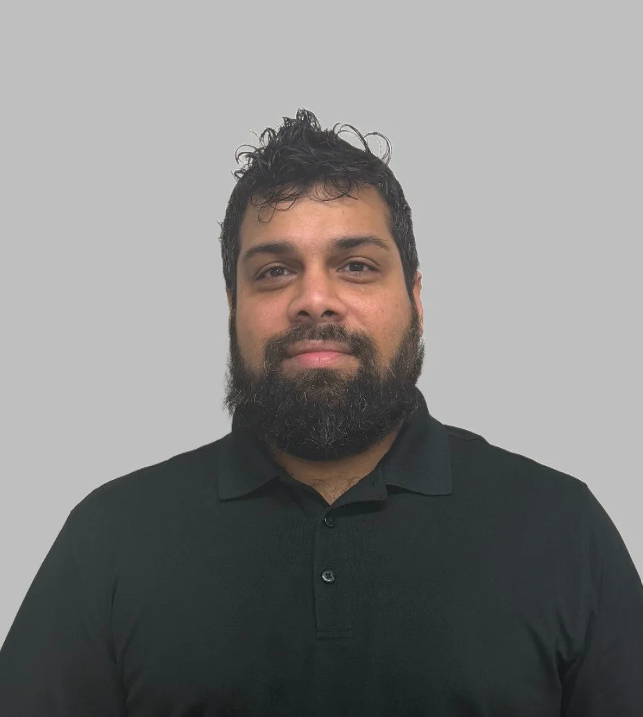 Headshot of a man with dark hair and a beard, wearing a black collared shirt, against a plain gray background.