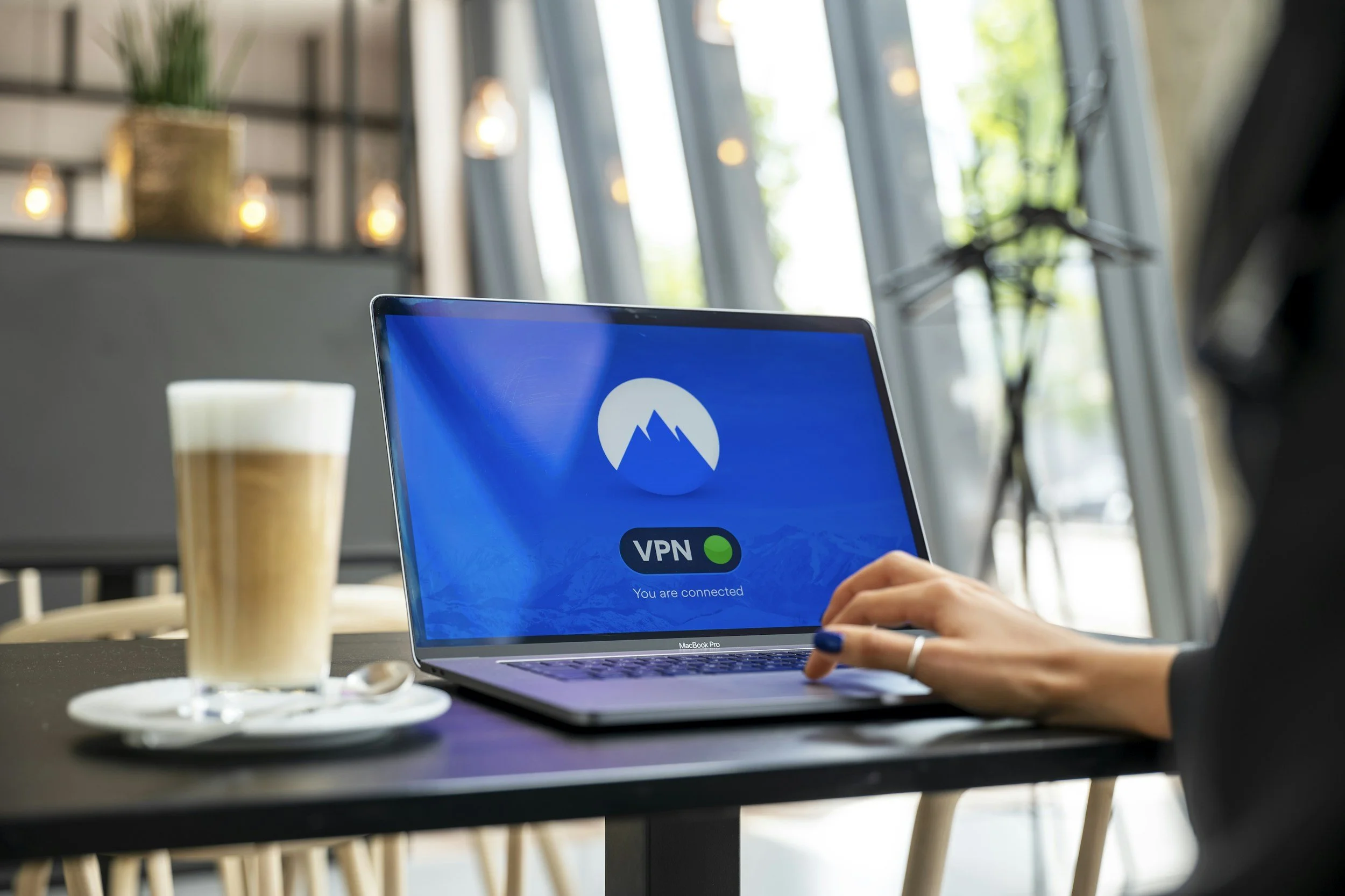 MacBook Pro displaying "VPN - You are connected" screen in a cafe setting, with a hand on the keyboard and a latte on the table.