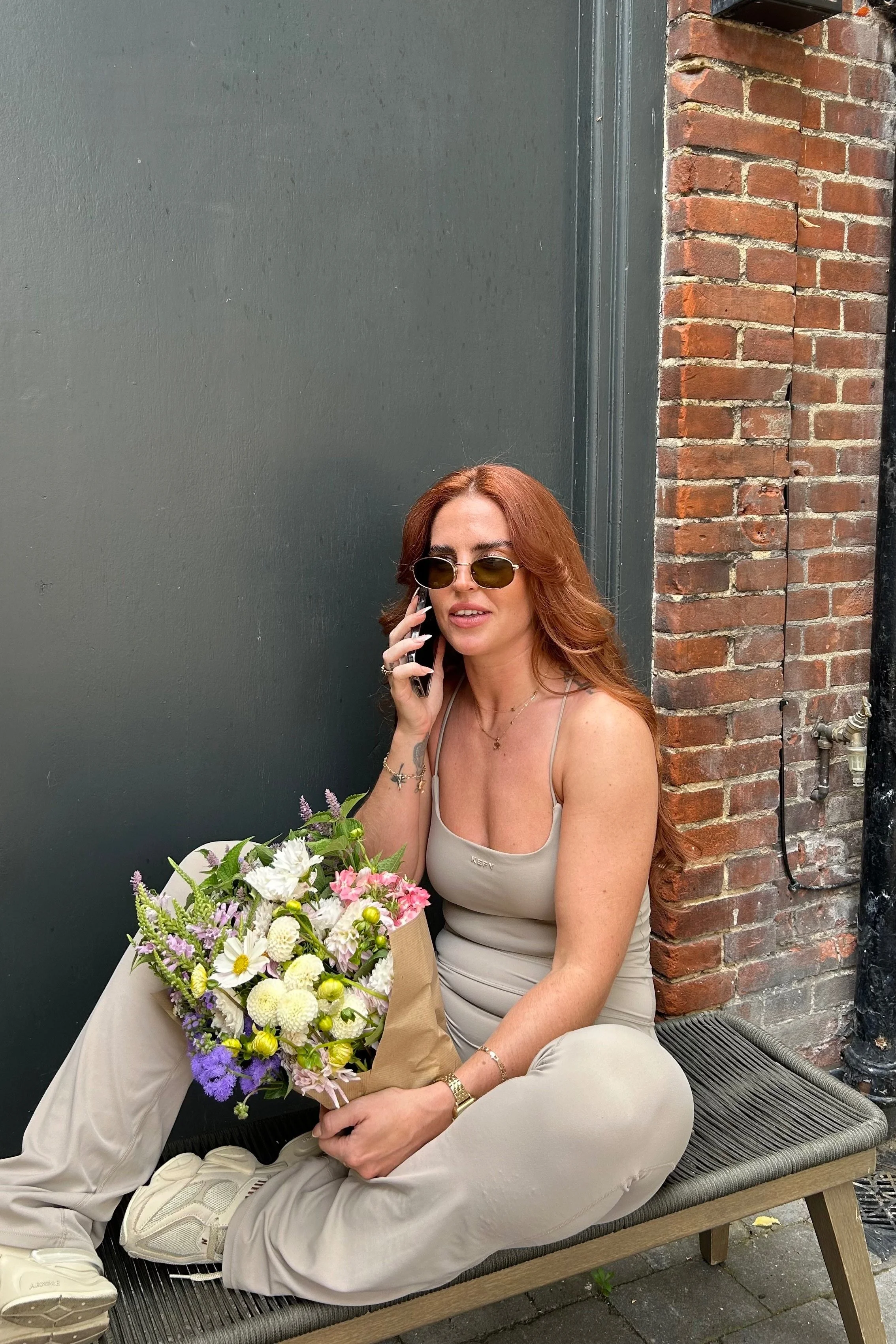 A woman with red hair, sunglasses, and a beige outfit sitting on a bench outside, talking on a cellphone, with a bouquet of flowers on her lap.