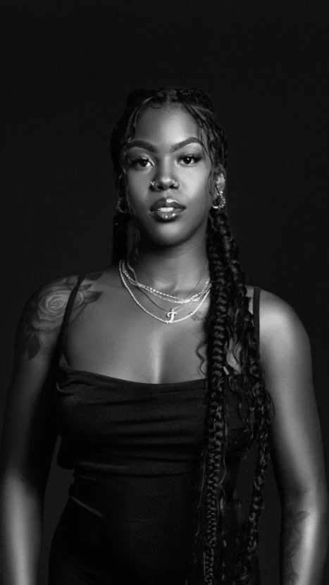 A black-and-white portrait of a young woman with long braided hair, wearing a sleeveless top, layered necklaces, and visible tattoos on her arm.