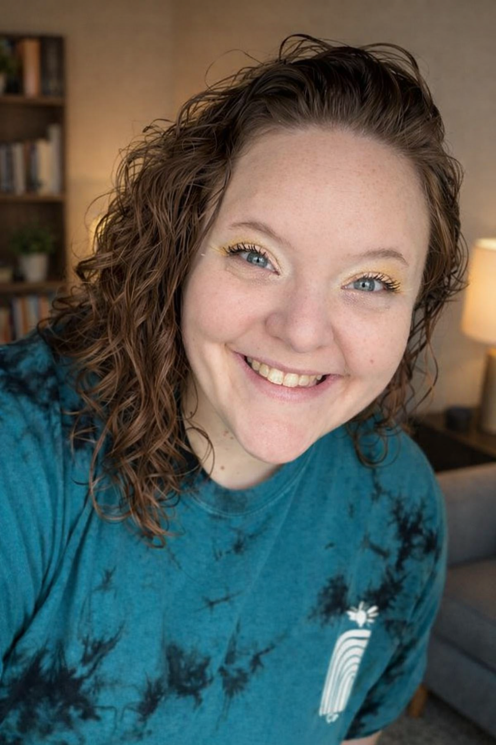 Close-up of a woman with curly brown hair smiling with teeth showing, wearing a gray shirt.