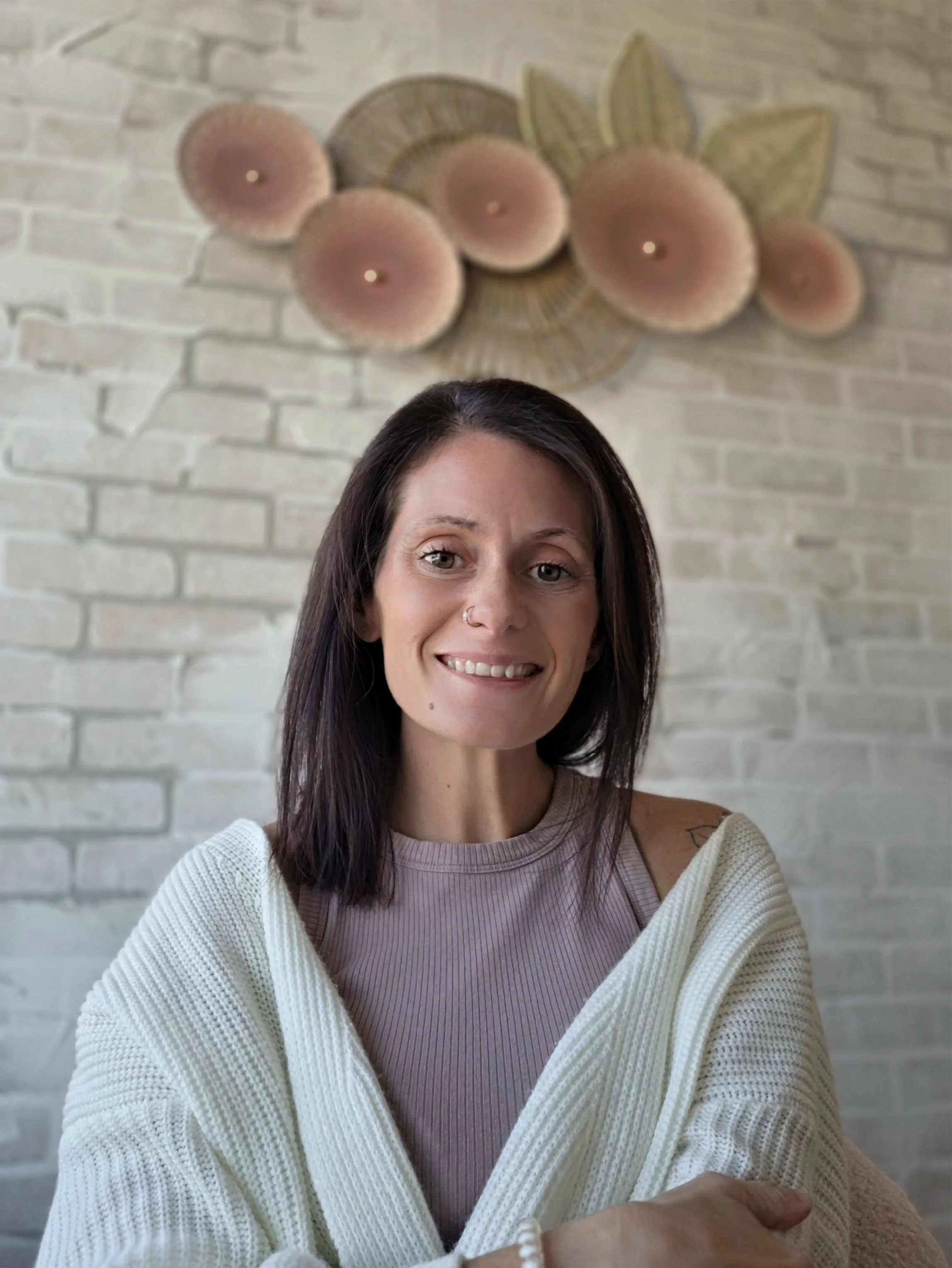 A smiling woman with brown wavy hair, light makeup, a nose piercing, and a small dot on her chin, wearing a beige knit turtleneck sweater.