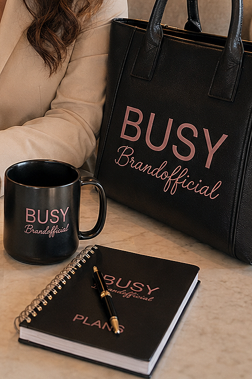 Black coffee mug, black handbag with pink text "BUSY Brandofficial," spiral-bound planner with pink text "BUSY Brandofficial PLANS," and a black pen, all on a white marble surface.
