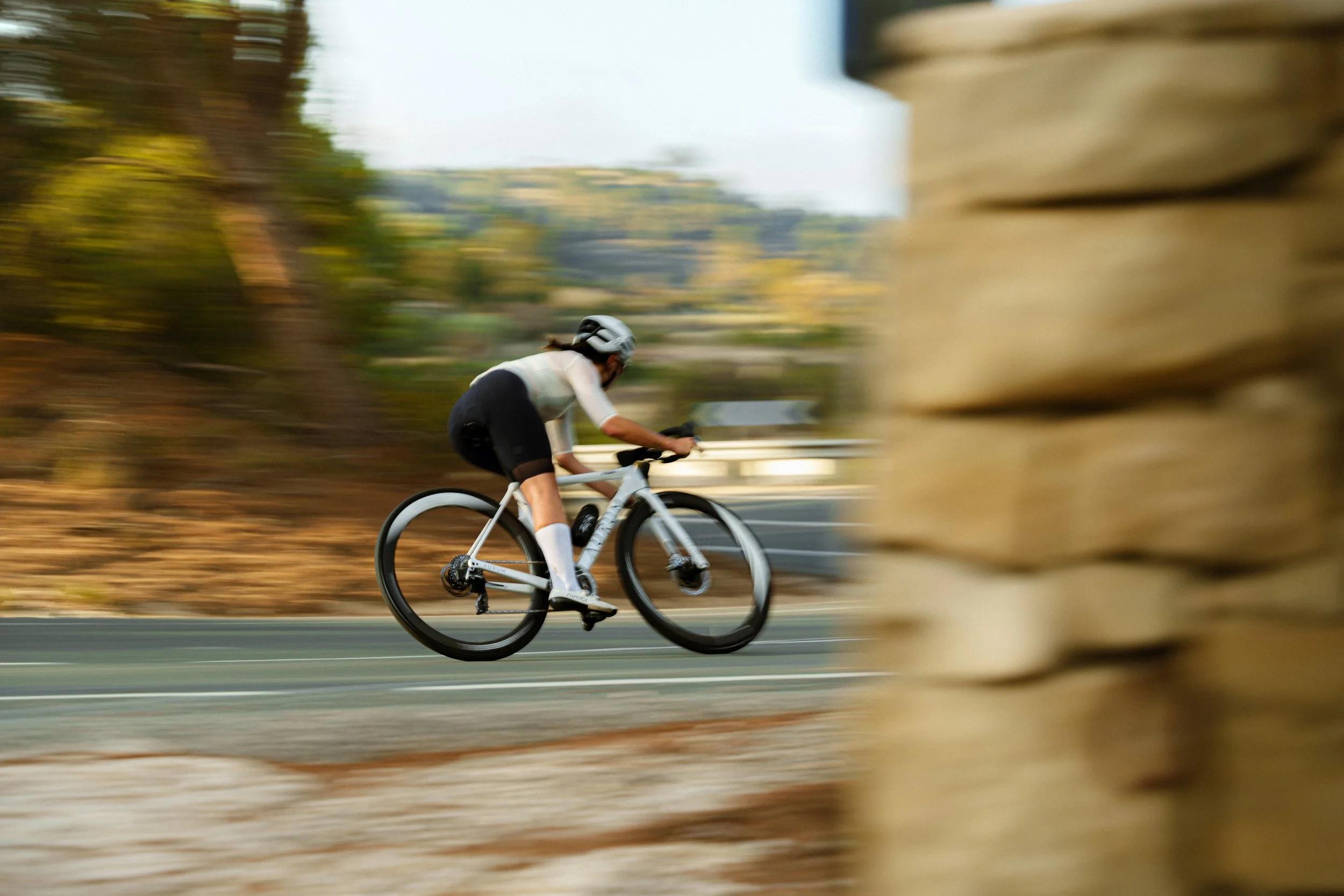 Cyclist in motion on a road bike, passing through a blurred outdoor environment, with a stone wall partially visible in the foreground.