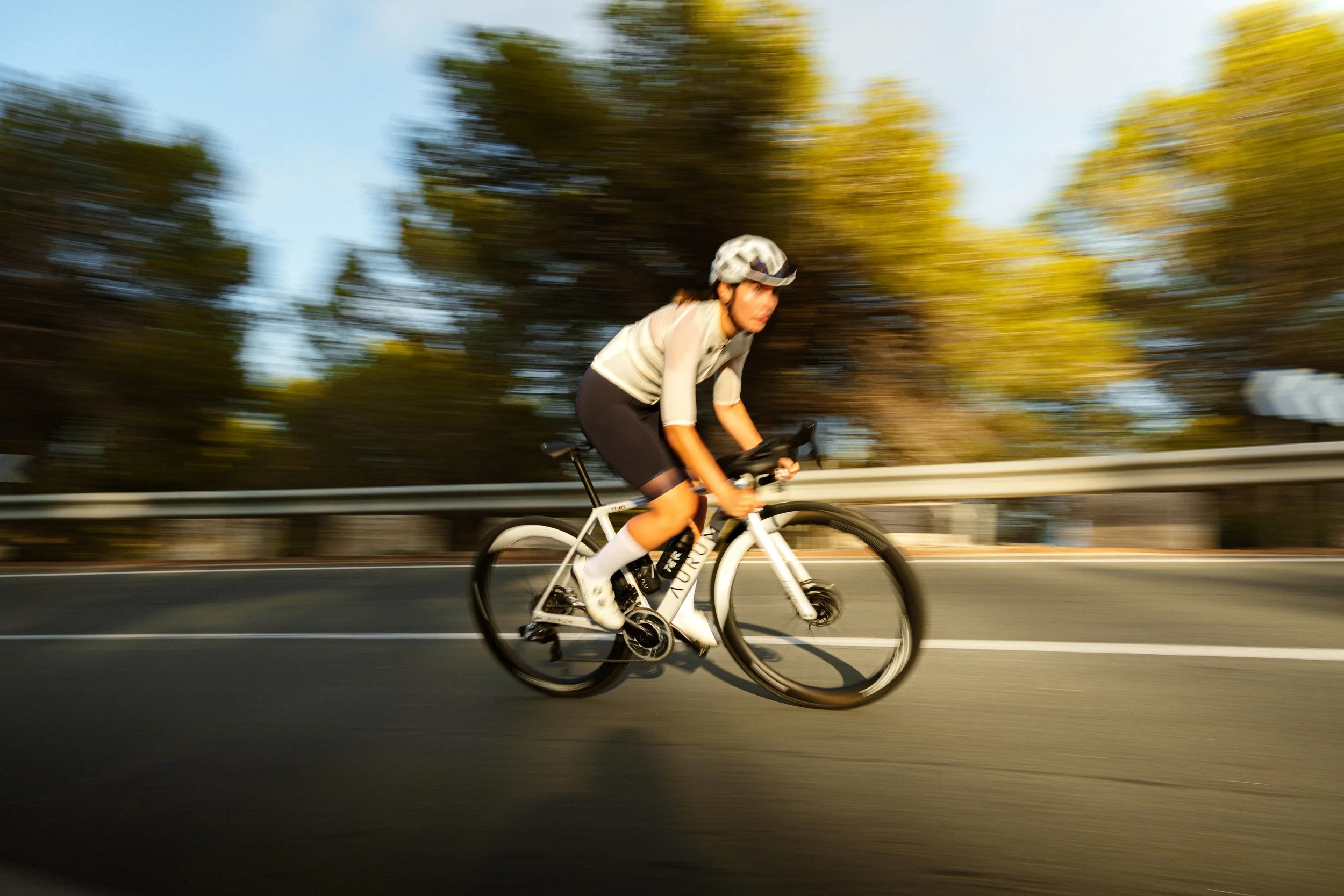 Cyclist on road bike riding fast on a road with blurred background of trees and guardrail.