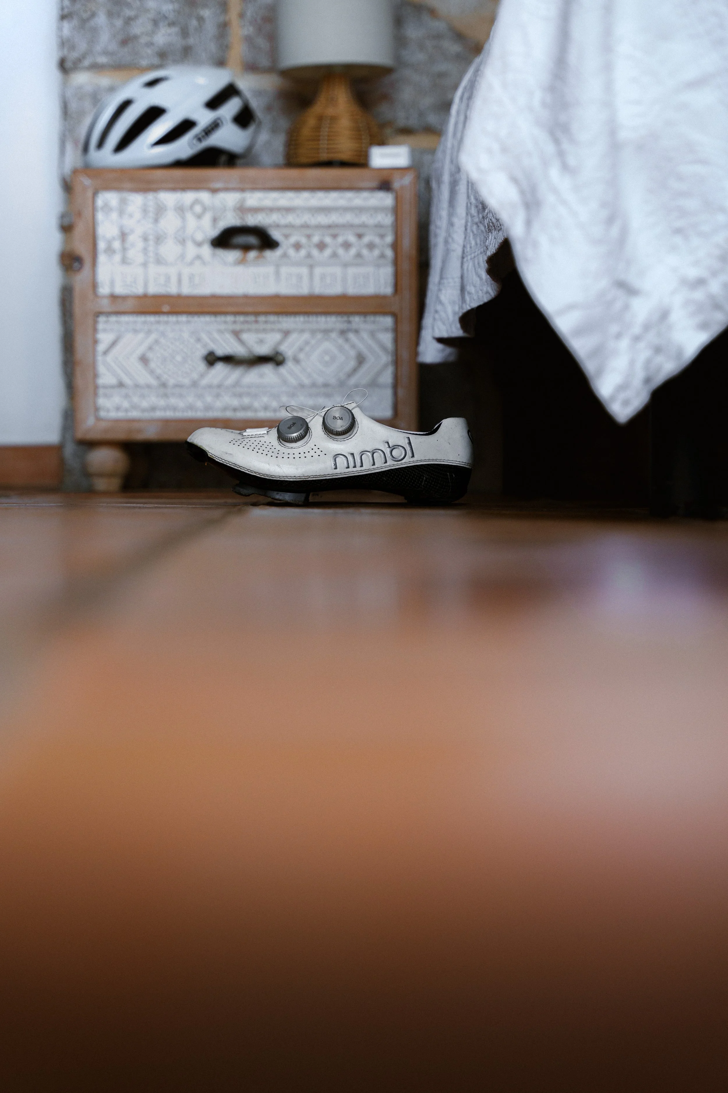White cycling shoe on the floor near a wooden nightstand with a bicycle helmet on top.