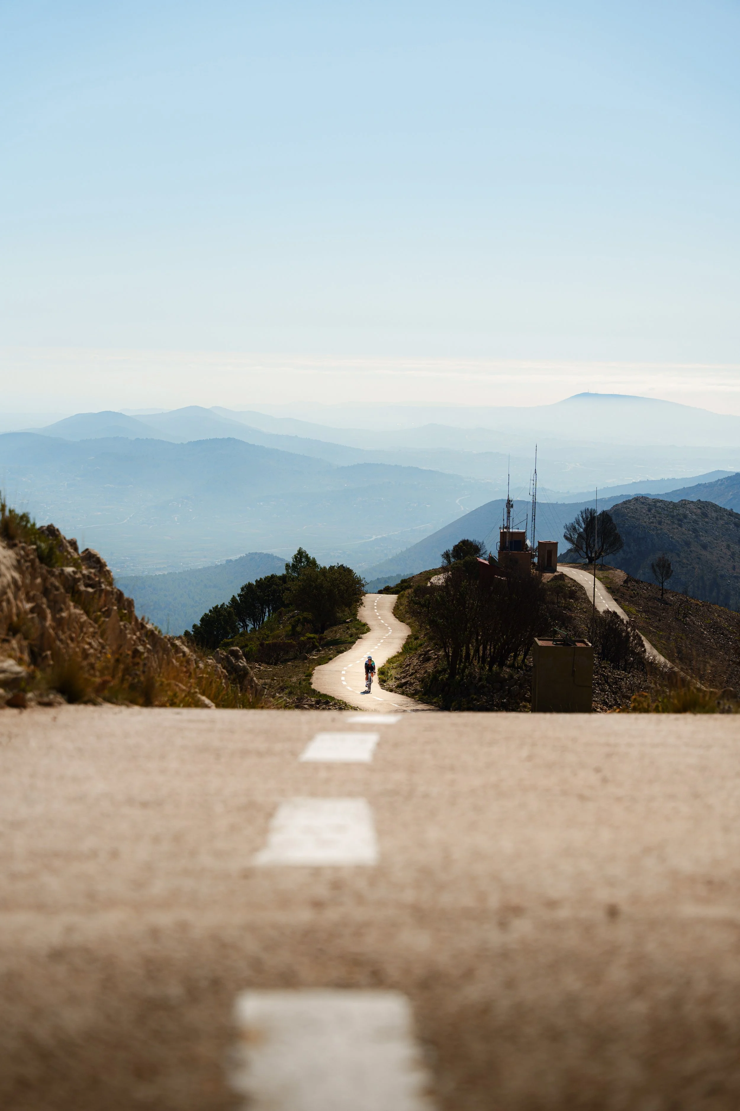 A cyclist on a winding mountain road with a distant view of mountains and a blue sky.