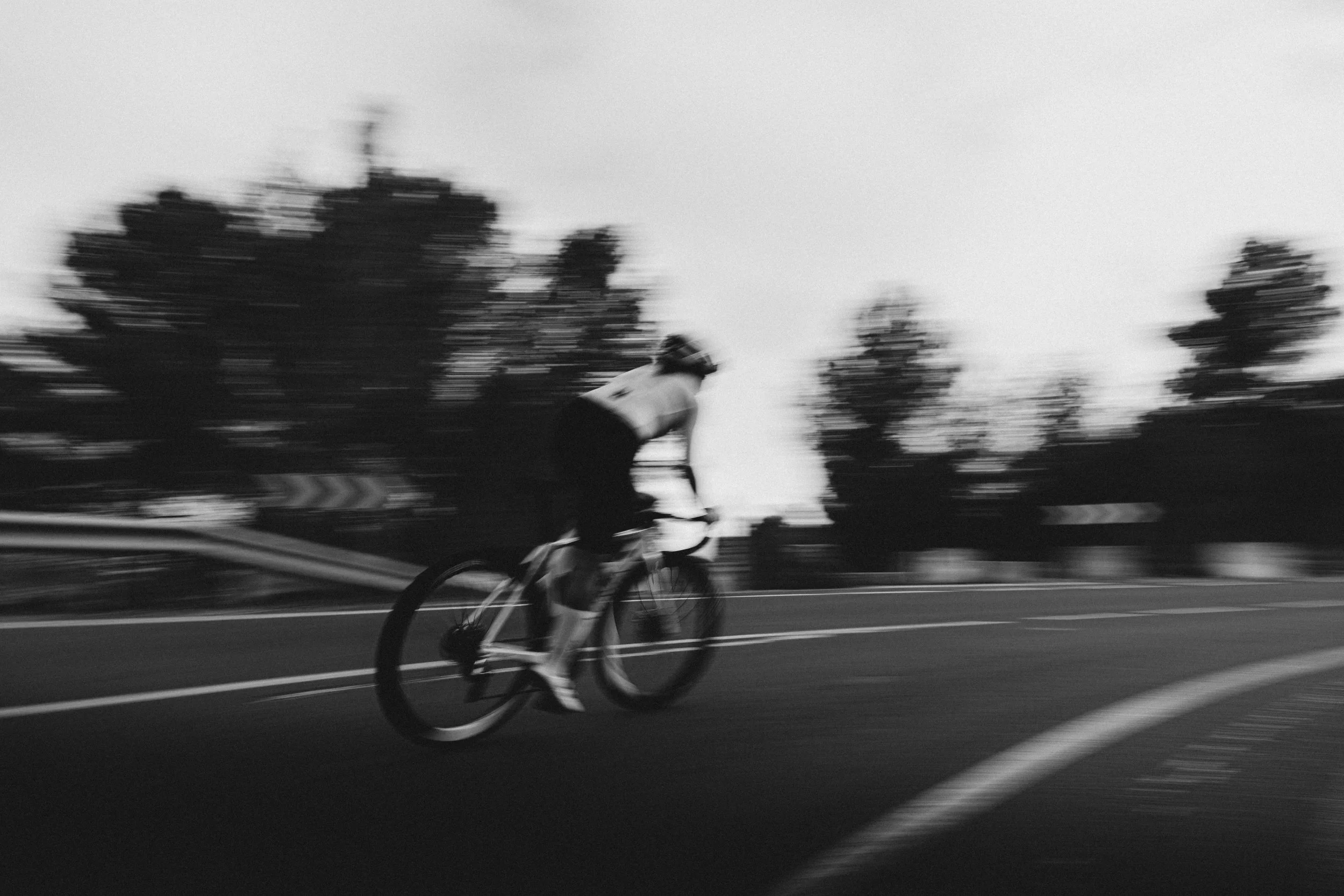 Black and white photo of a cyclist on a road, motion blur effect, trees in background.