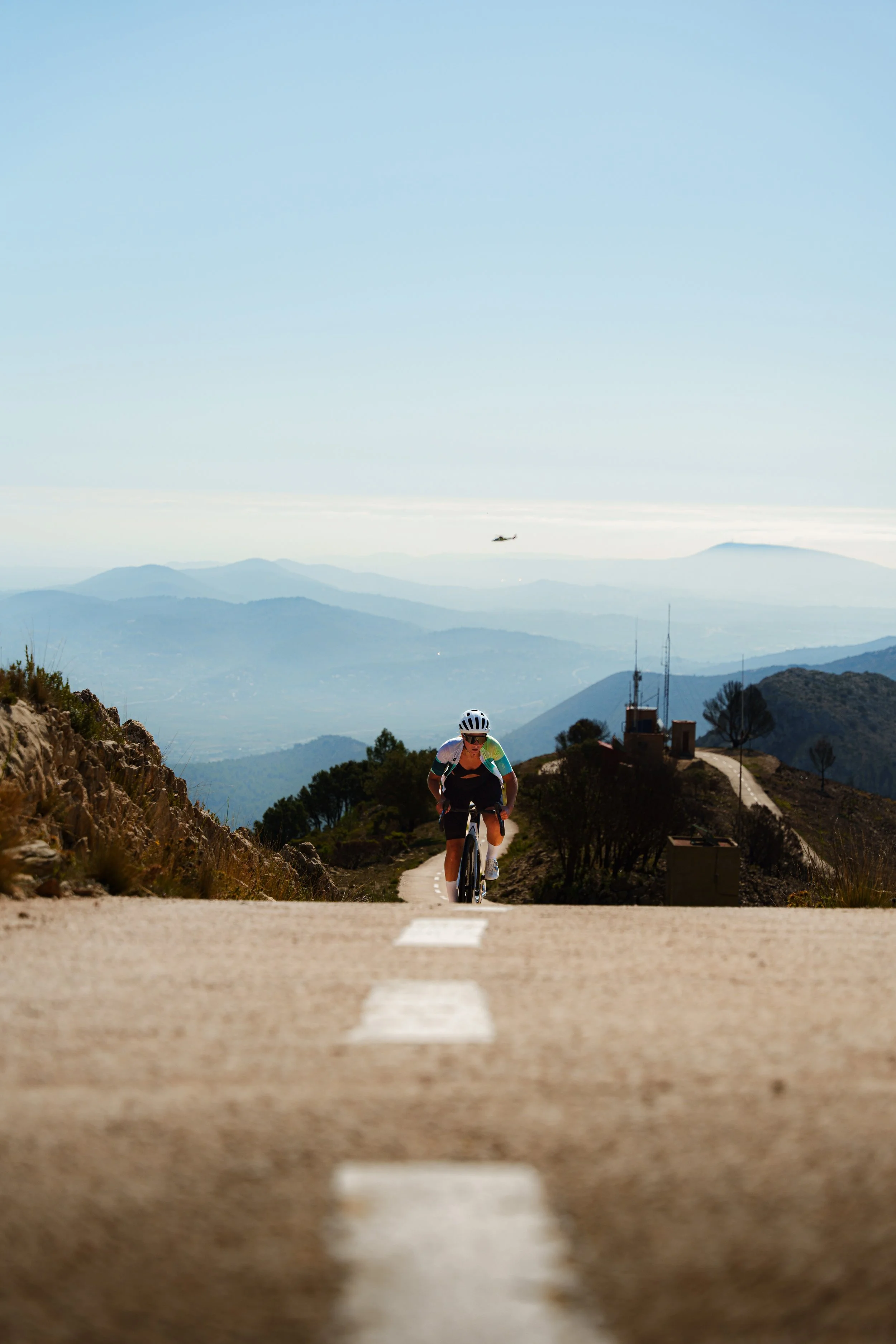 Cyclist riding uphill on a winding mountain road, with distant hills and a clear sky in the background. A helicopter flies in the sky.