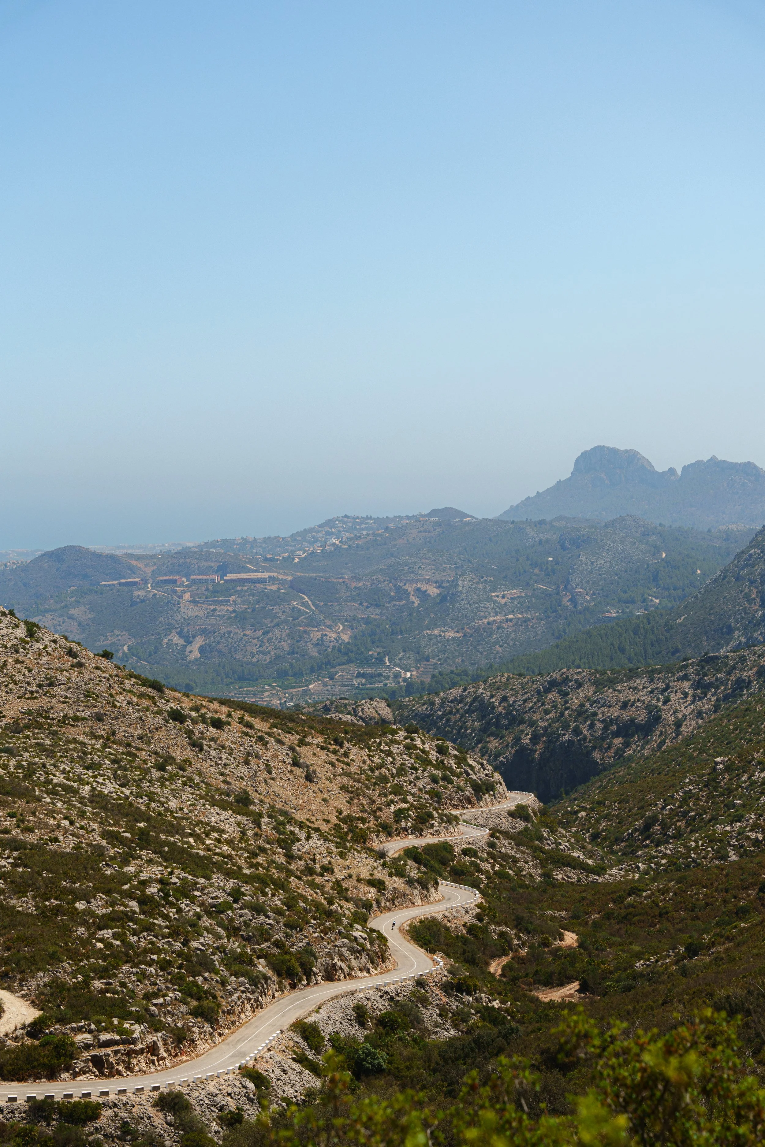 Winding road through a rocky mountainous landscape with distant hills and a clear blue sky.