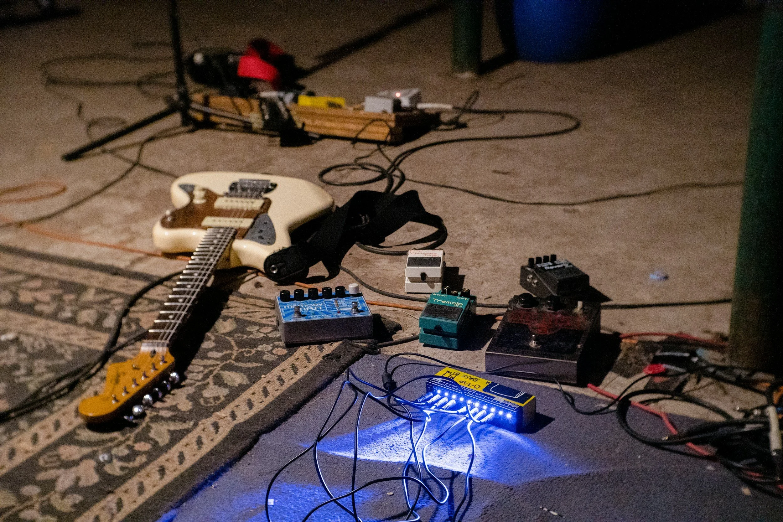Guitar and various guitar effect pedals and gear arranged on a carpeted floor, with cables connected to a blue-lit effects box.