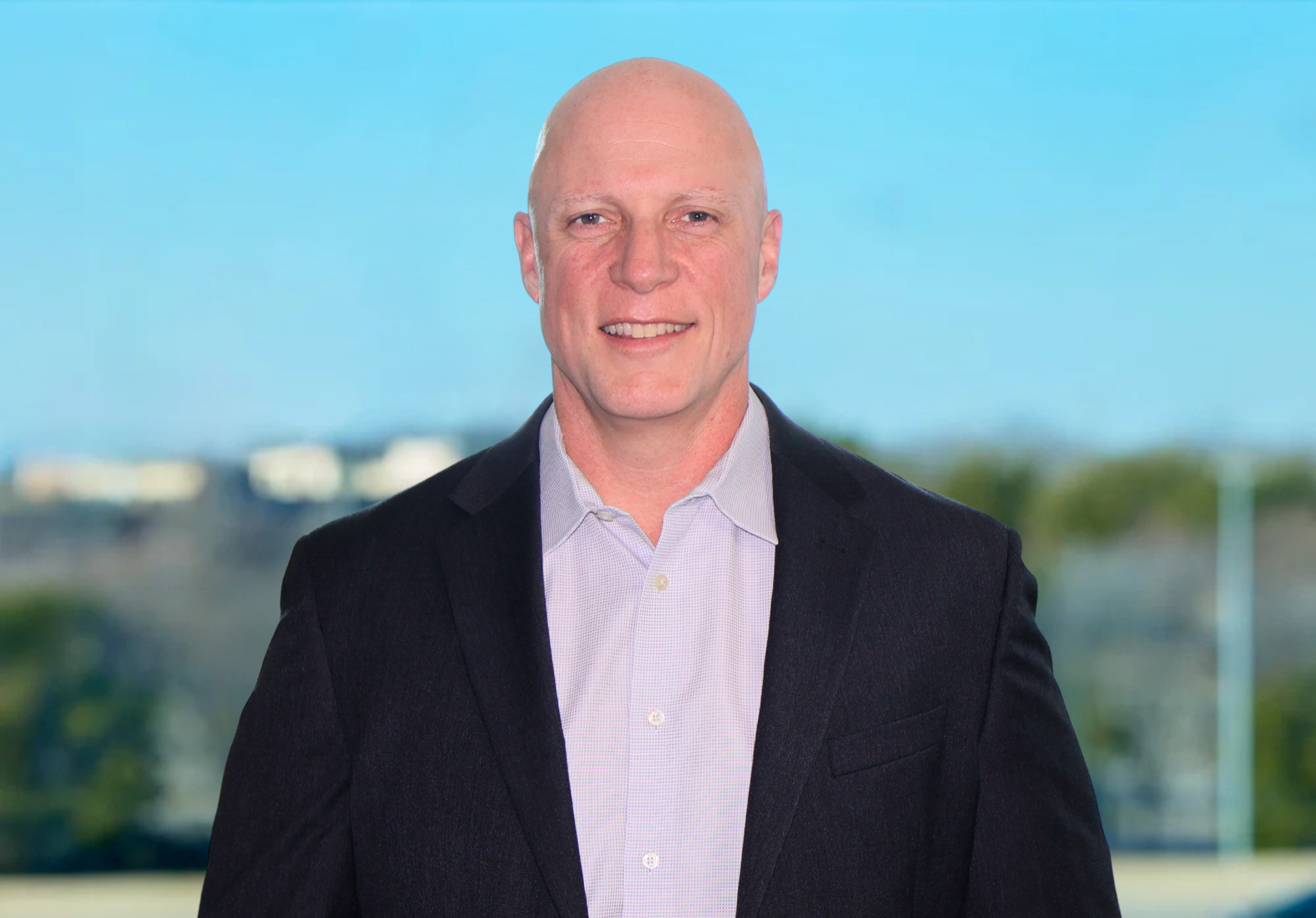 Portrait of a bald man wearing a dark button-up shirt smiling in front of a blurred outdoor background with blue sky.