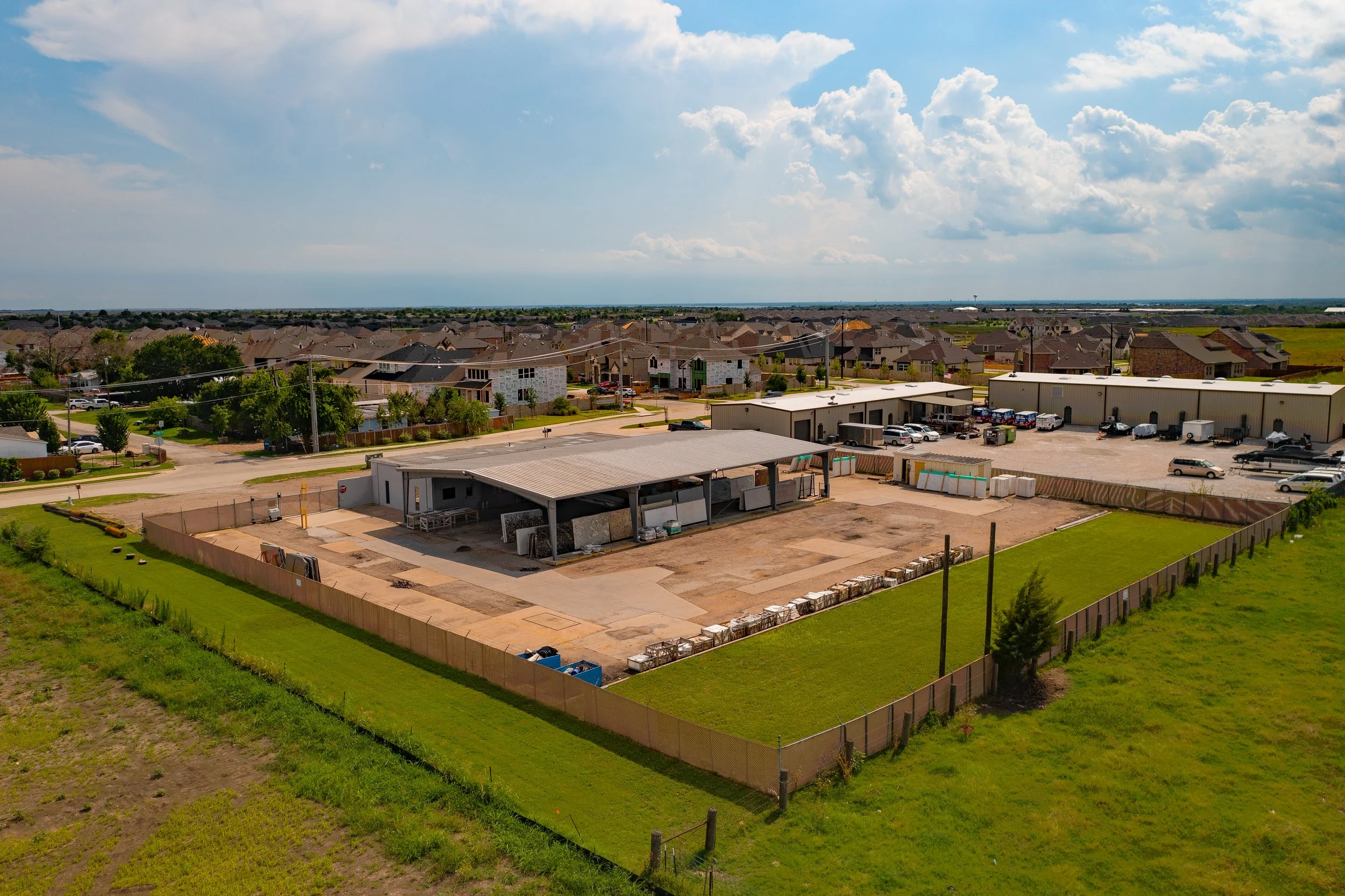 Aerial view of a construction site with a partially built structure, surrounded by a fence, adjacent to a parking lot with several parked vehicles, in a suburban neighborhood with new houses in the background.
