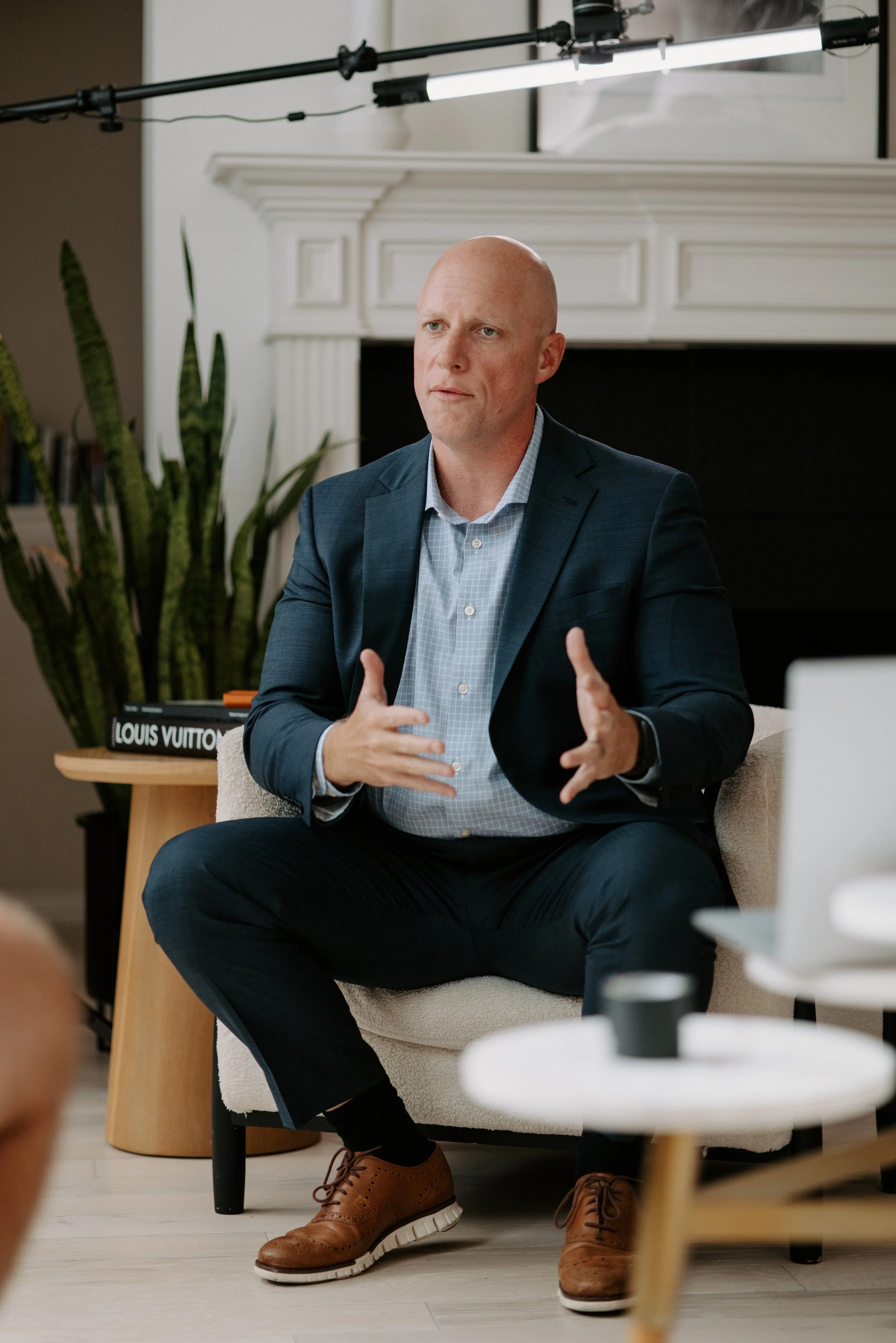 A man in a dark suit and blue dress shirt sitting on a beige chair, speaking with hand gestures during an indoor meeting or interview. Background includes large plants and a white fireplace, with recording lighting overhead.