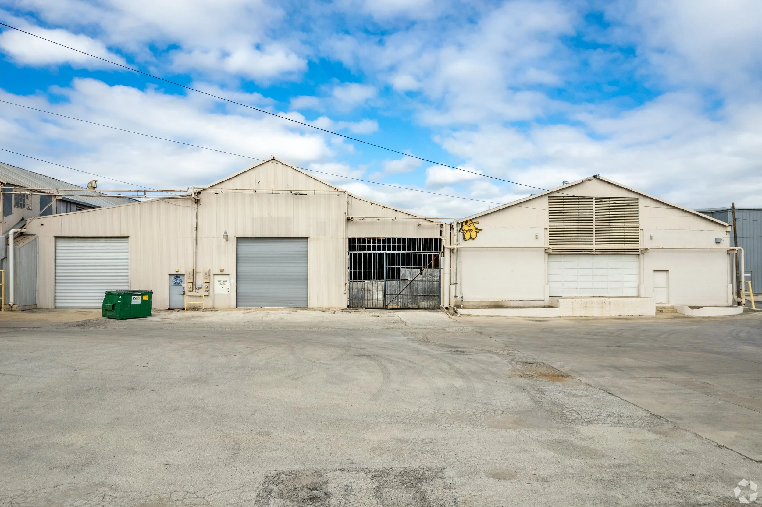 Industrial warehouses with metal siding, roll-up doors, and a gate, under a partly cloudy sky.