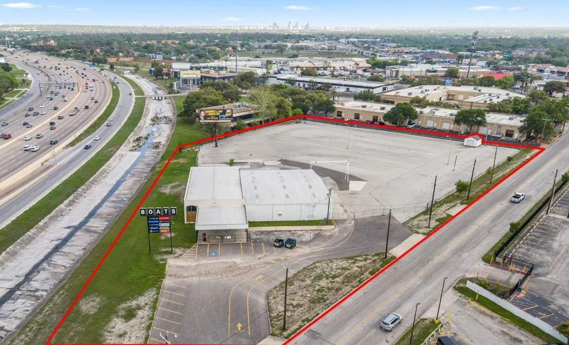 Aerial view of a commercial property outlined in red, featuring a building with a boat dealership sign, a large parking lot, and a basketball court, cityscape in the background.