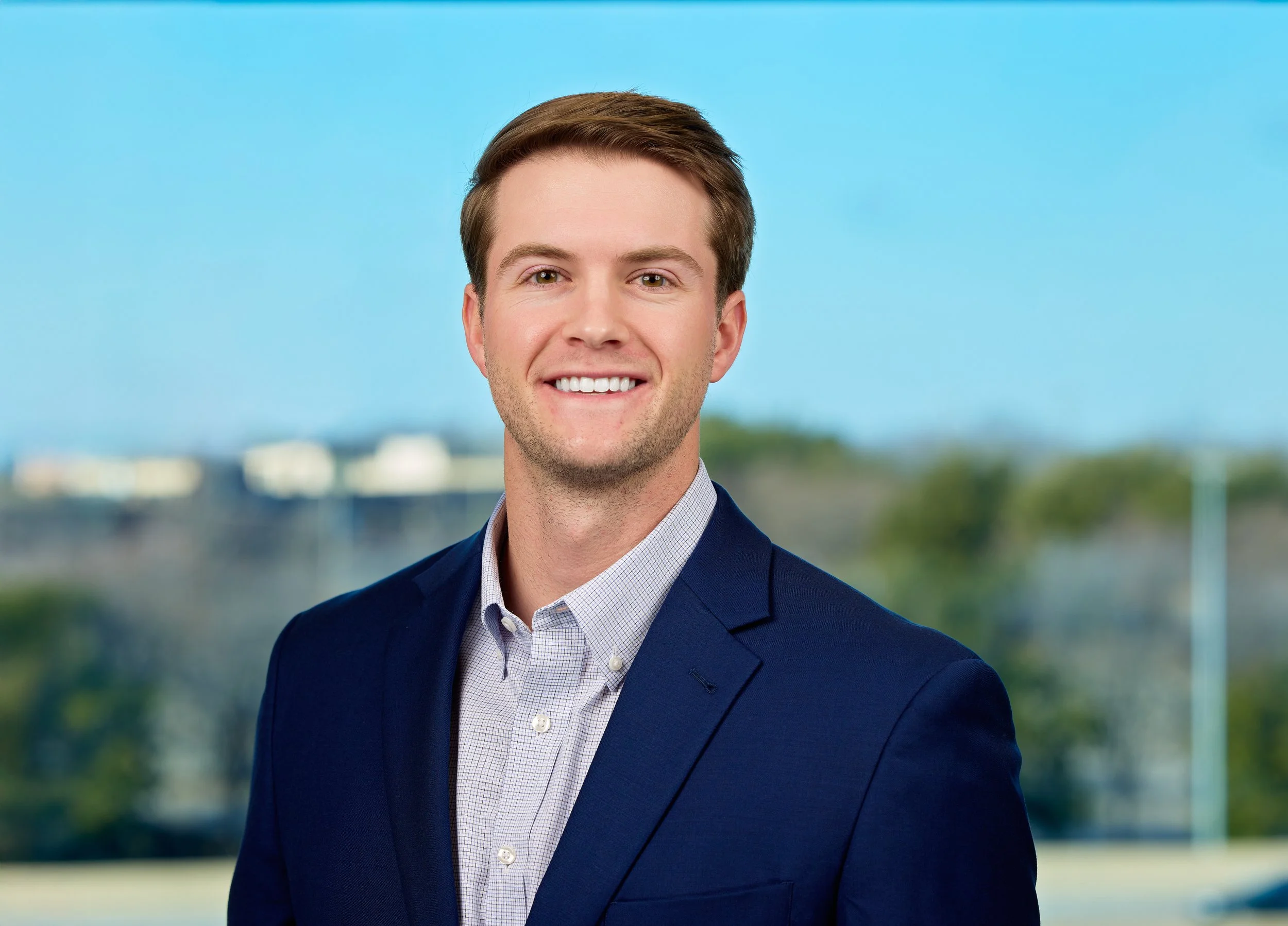 Headshot of a young man with brown hair, smiling, wearing a navy blazer and a light checkered shirt, standing outdoors with a blurred cityscape and blue sky in the background.