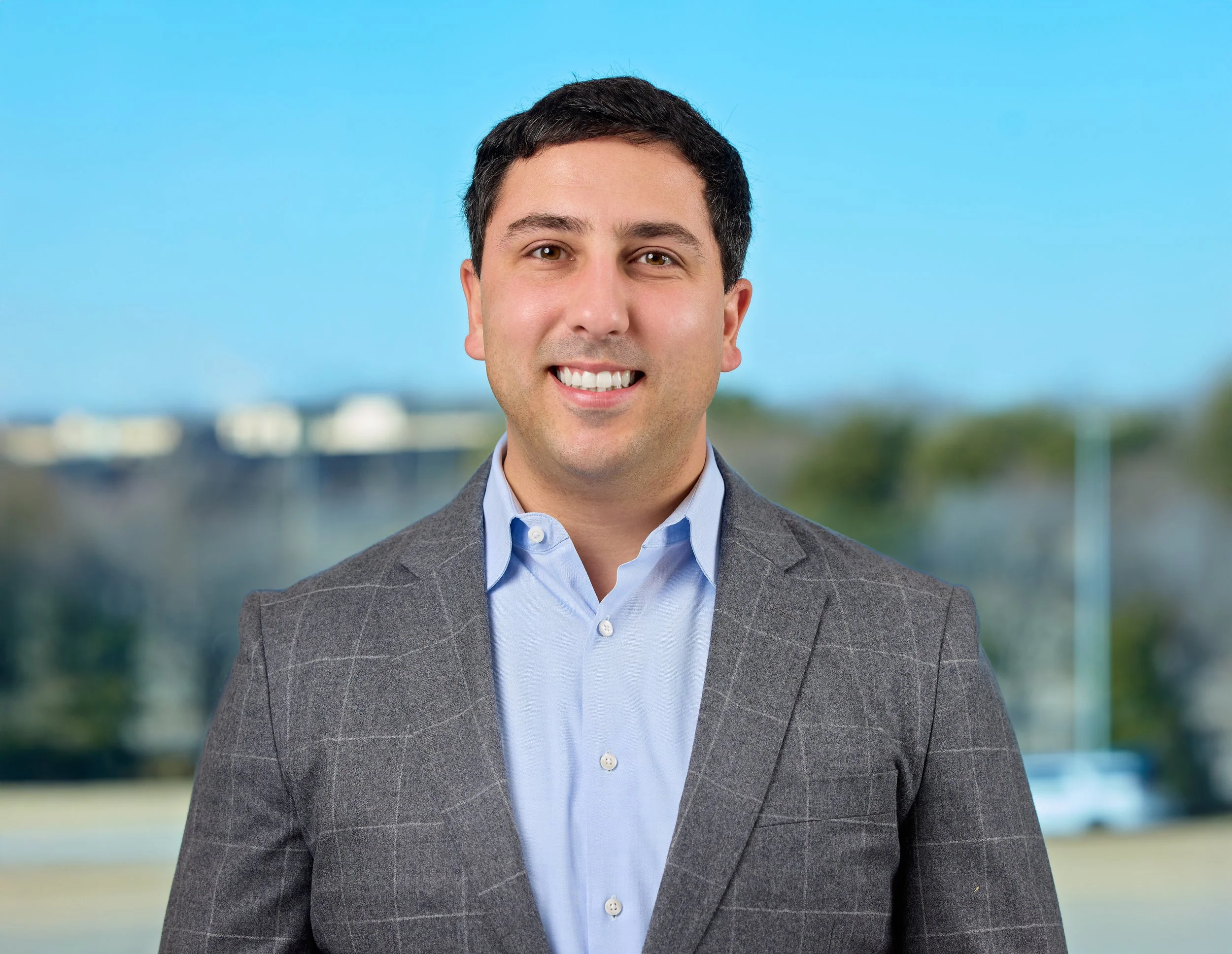 Portrait of a smiling man in a gray checkered blazer and light blue shirt, standing outdoors with blue sky and blurred buildings in the background.