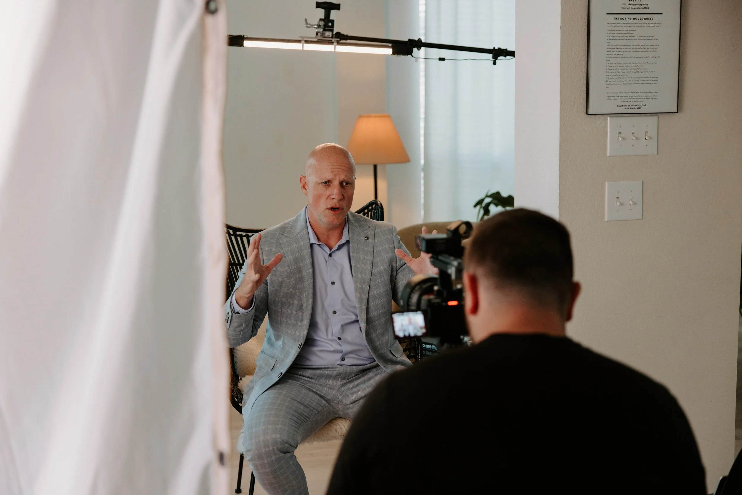 A man in a light gray suit is speaking during an interview or recording session in a home setting, as seen through a partially closed white curtain. A camera operator is filming him, and the scene is set with a background including a floor lamp, some chairs, and houseplants.