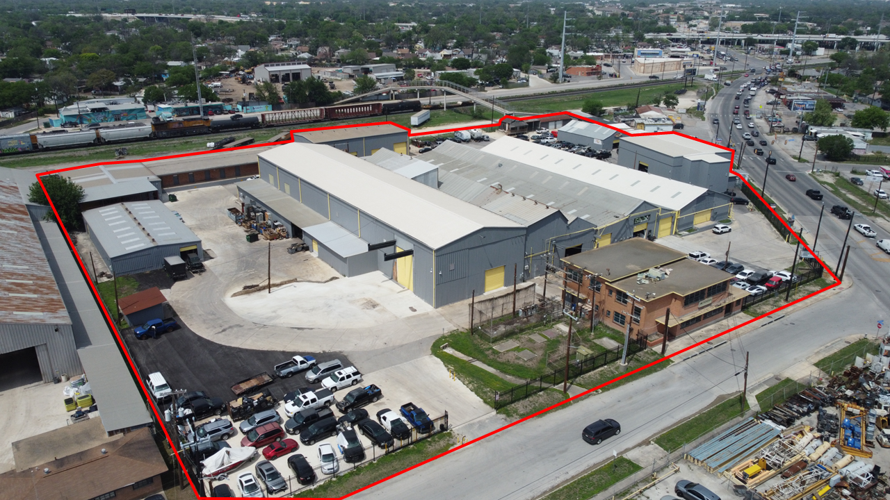 Aerial view of a large industrial warehouse complex outlined in red, with parking lots, a small brick building, and surrounding streets with cars in an urban area.