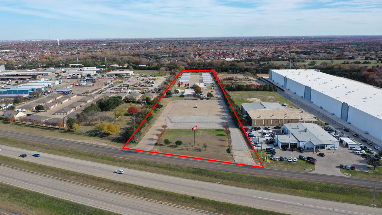 Aerial view of a commercial land lot outlined in red, with surrounding warehouses, parking lots, highway, and suburban neighborhood in the background.