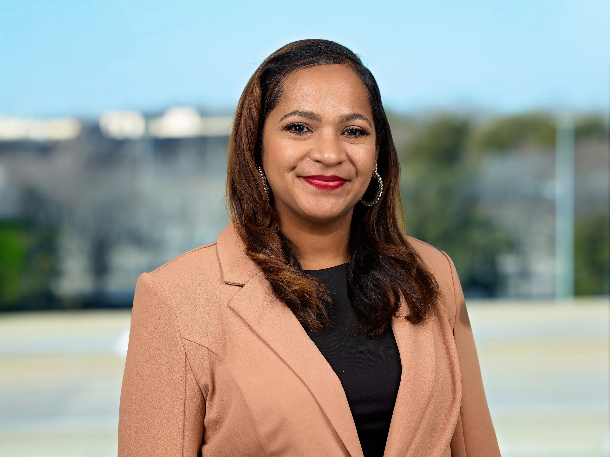 Headshot of a confident woman with medium skin tone, brown wavy hair, wearing a light brown blazer over a black top, standing outdoors with a blurred cityscape background.