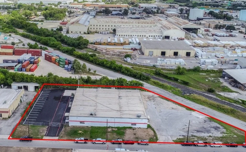 Aerial view of an industrial area showing a single-story building with a parking lot, surrounded by a red boundary line, adjacent to train tracks, with warehouses, storage containers, and other industrial buildings in the background.