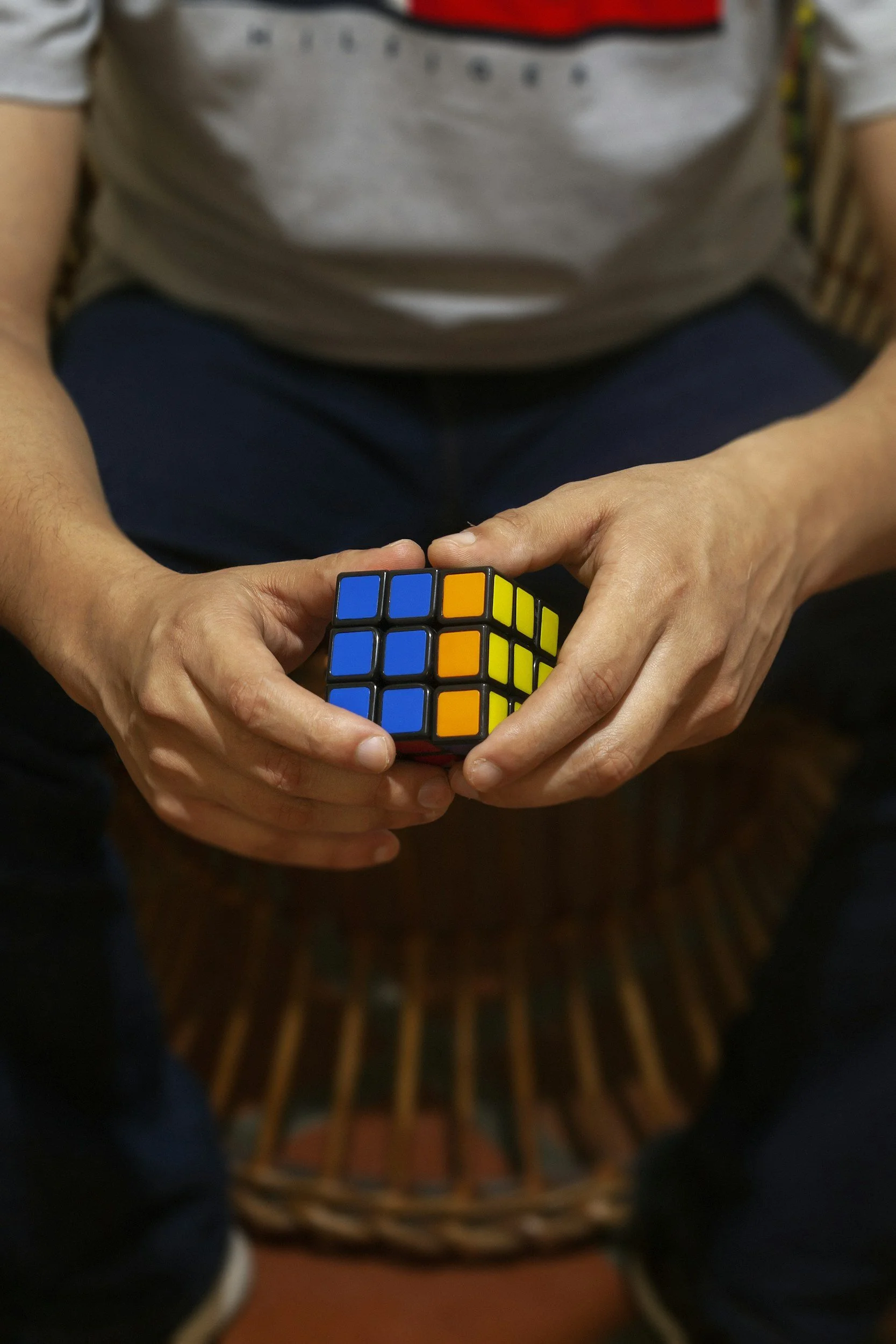 Person holding a Rubik's Cube with a colorful pattern, sitting in front of a woven wicker chair.